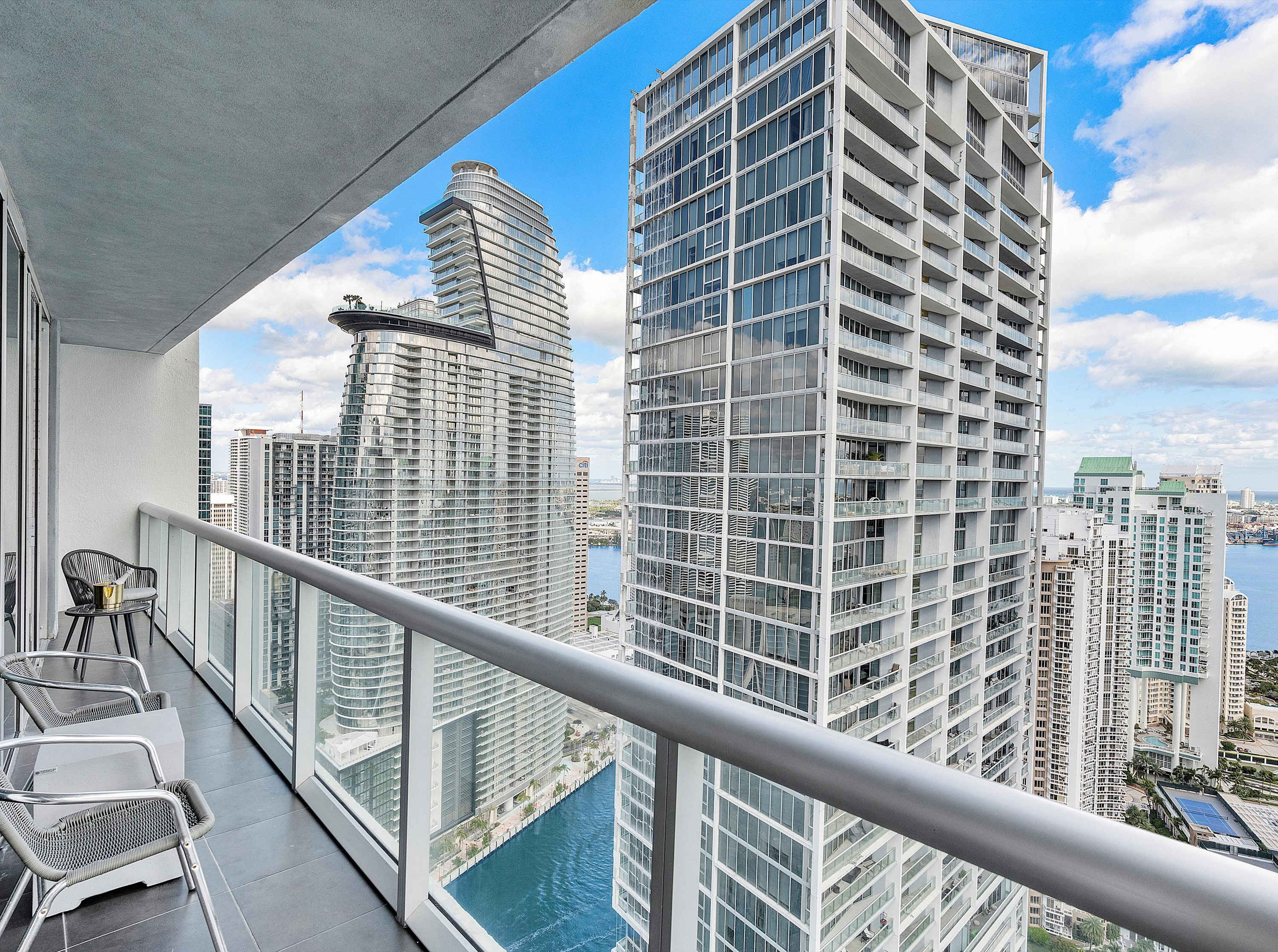 Outdoor balcony with modern grey chairs and a glass railing, offering a high-rise view of surrounding city buildings and a waterway below.