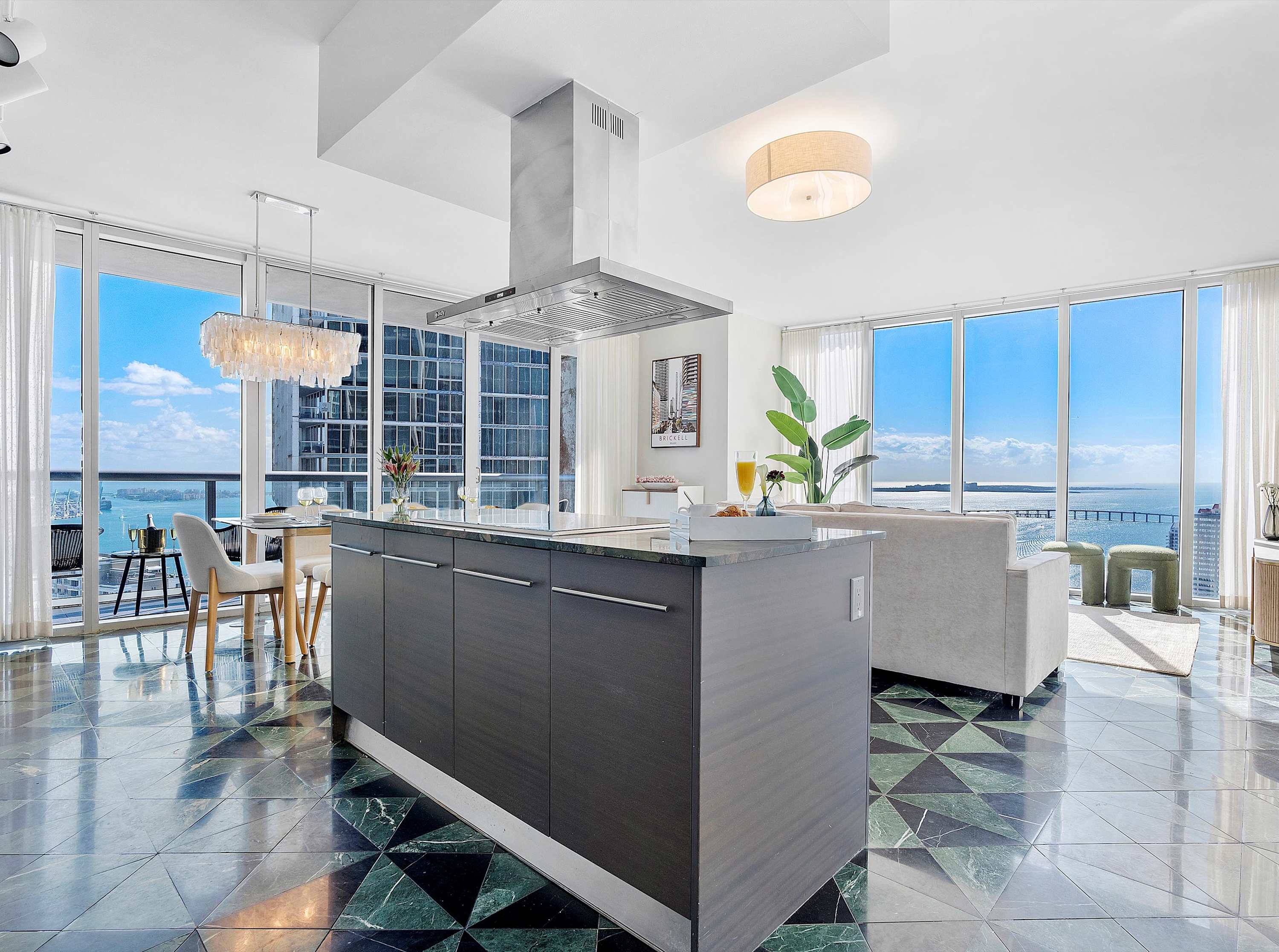 Open-concept kitchen and dining area with a large dark gray island, stainless steel appliances, and a glass dining table with white chairs, all overlooking the ocean through floor-to-ceiling windows, on a distinctive green and black geometric tile floor.