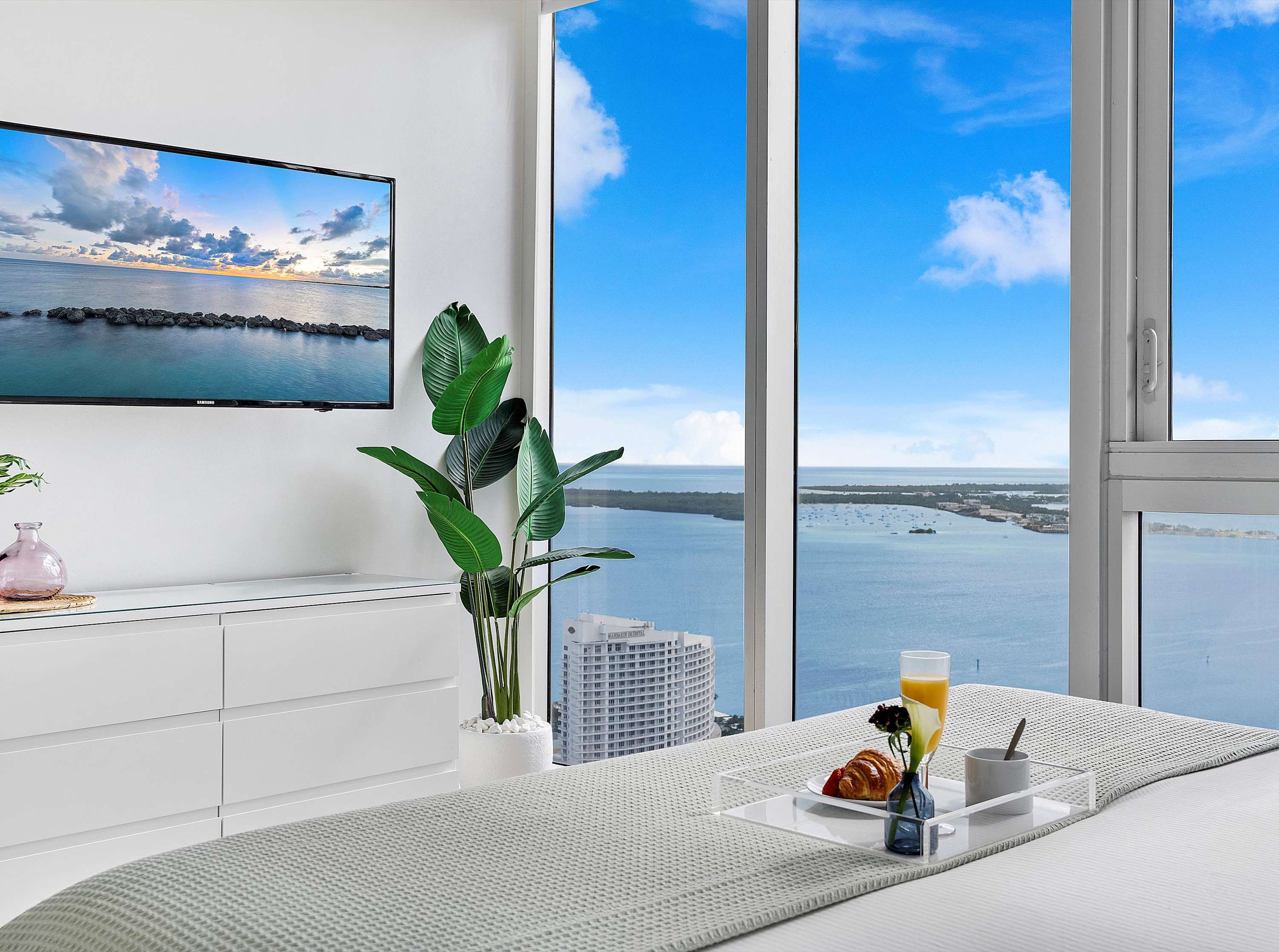 Bedroom with a large bed, a white dresser with a TV mounted above it, and floor-to-ceiling windows providing a stunning ocean view, featuring a breakfast tray on the bed and a potted plant.