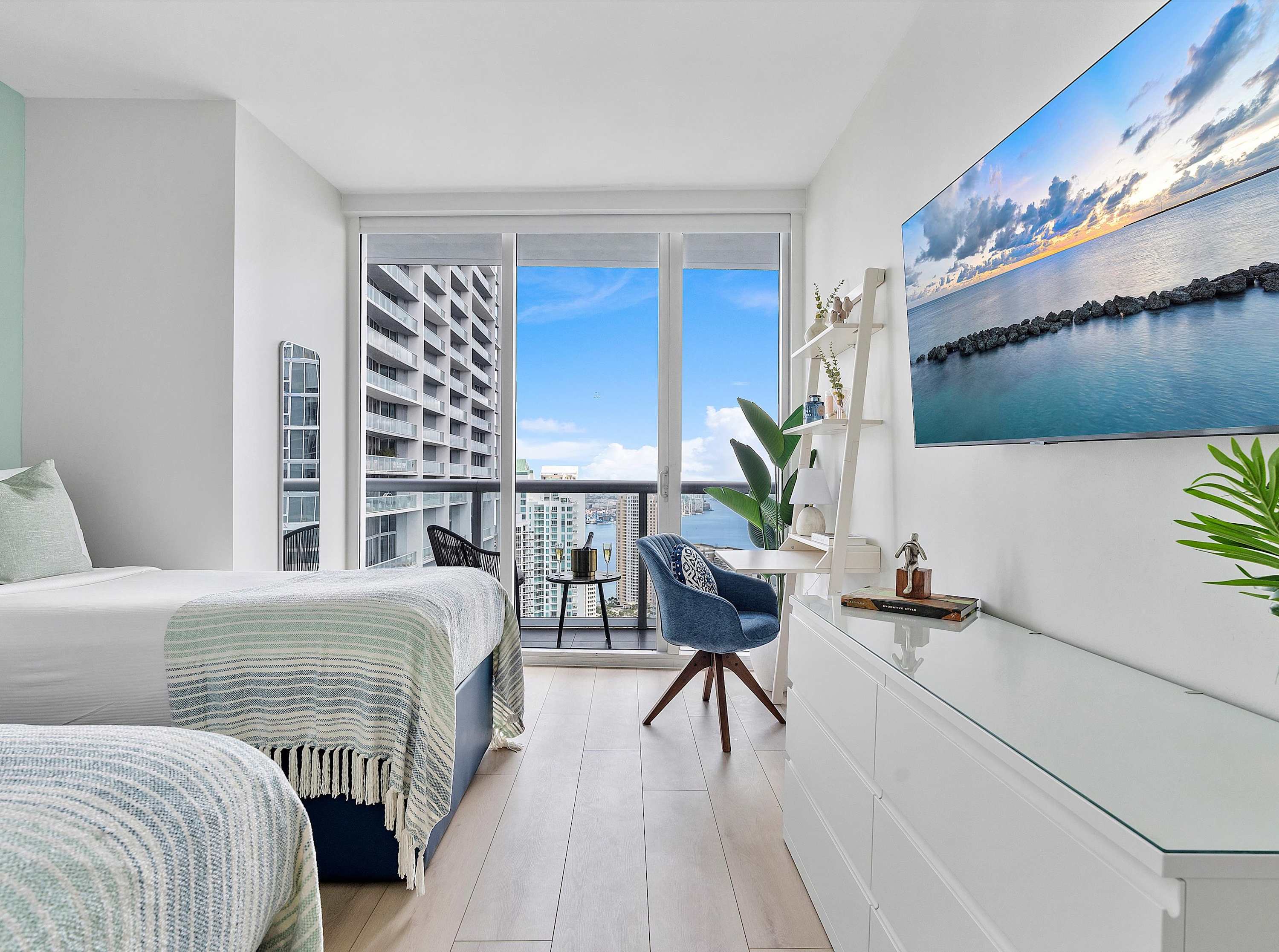 Bright bedroom with a white bed, light wood floors, a white desk with a blue chair, and a large TV mounted on the wall, offering expansive city and water views from a private balcony.