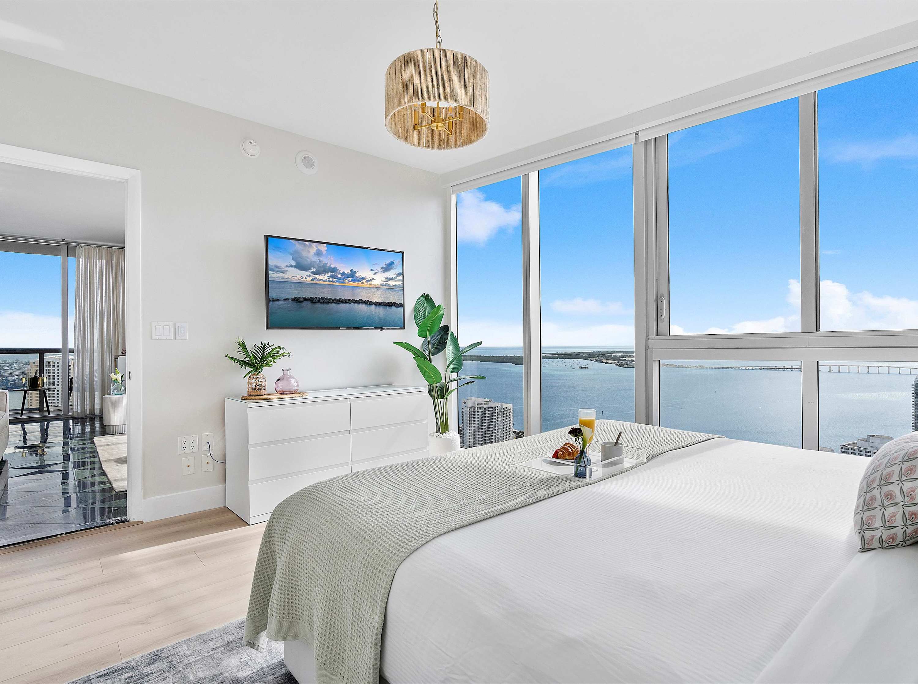 Bright master bedroom with a white bed, dresser, and wall-mounted TV, featuring floor-to-ceiling windows offering panoramic city and waterfront views.