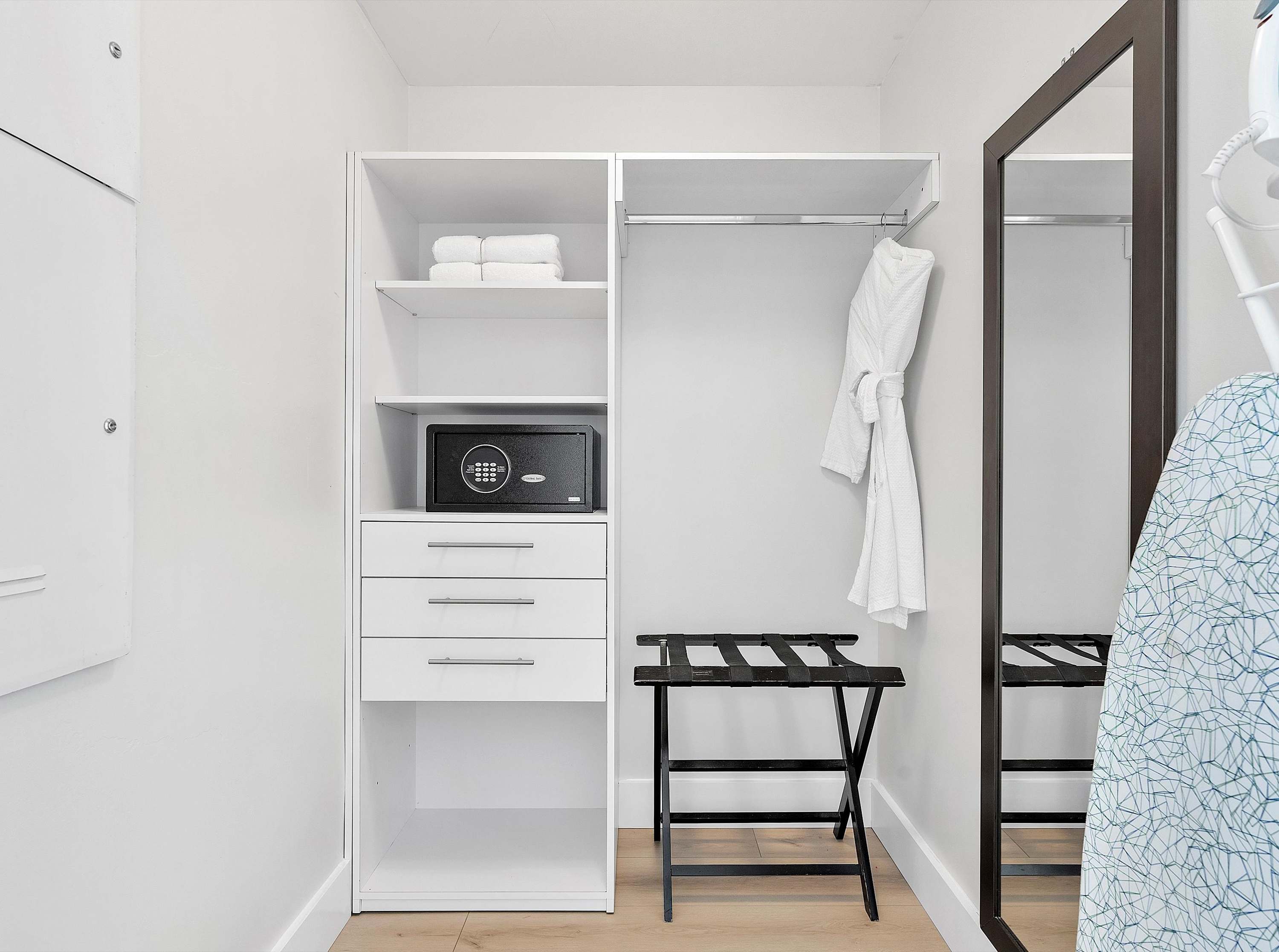 White built-in closet with shelves, drawers, a safe, and a hanging rod, accompanied by a black luggage rack and a full-length mirror.