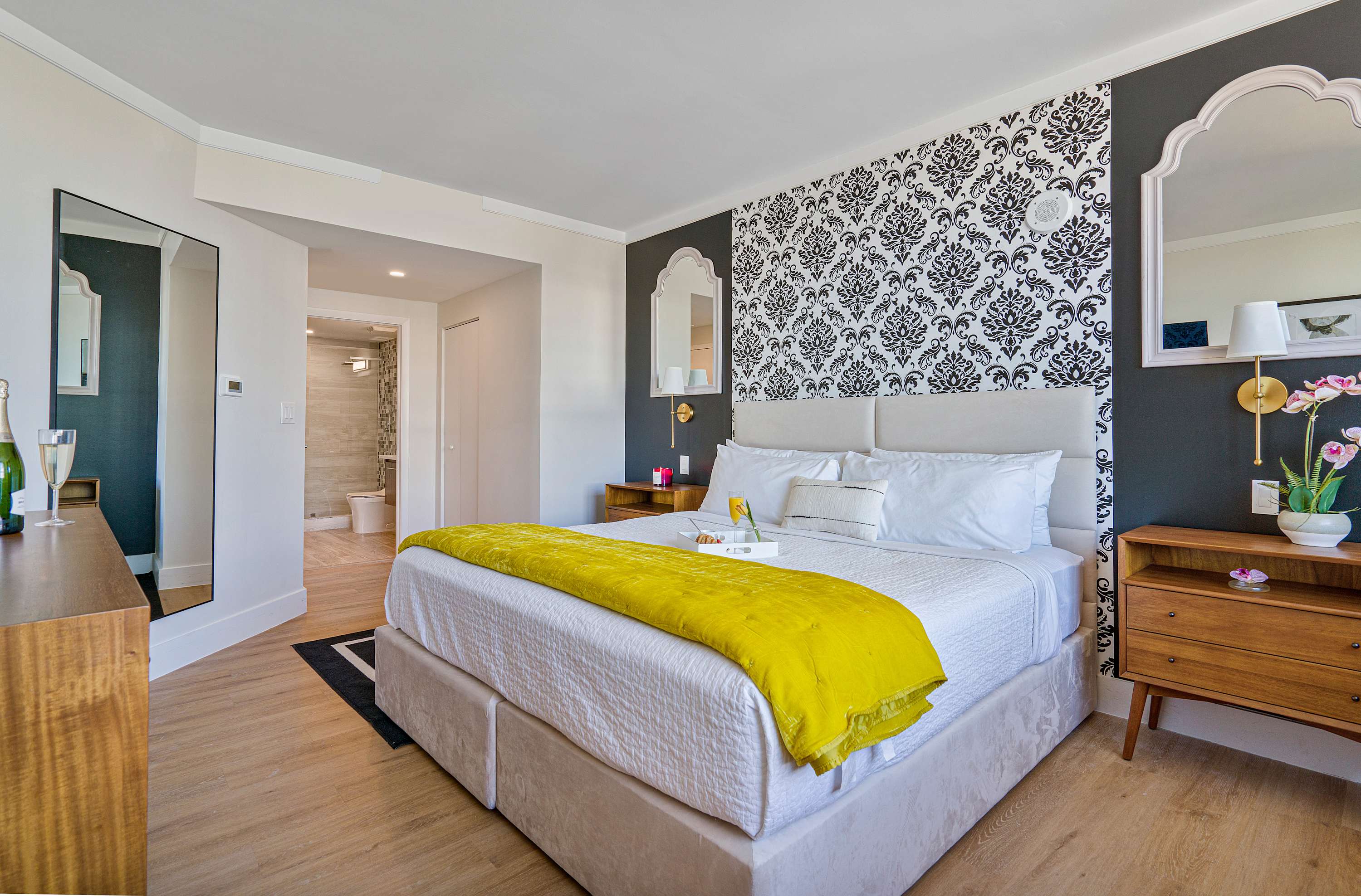 Elegant bedroom with a king-size bed, white headboard, and a striking black and white damask accent wall, featuring a full-length mirror and a wooden dresser, with a glimpse into the bathroom beyond.