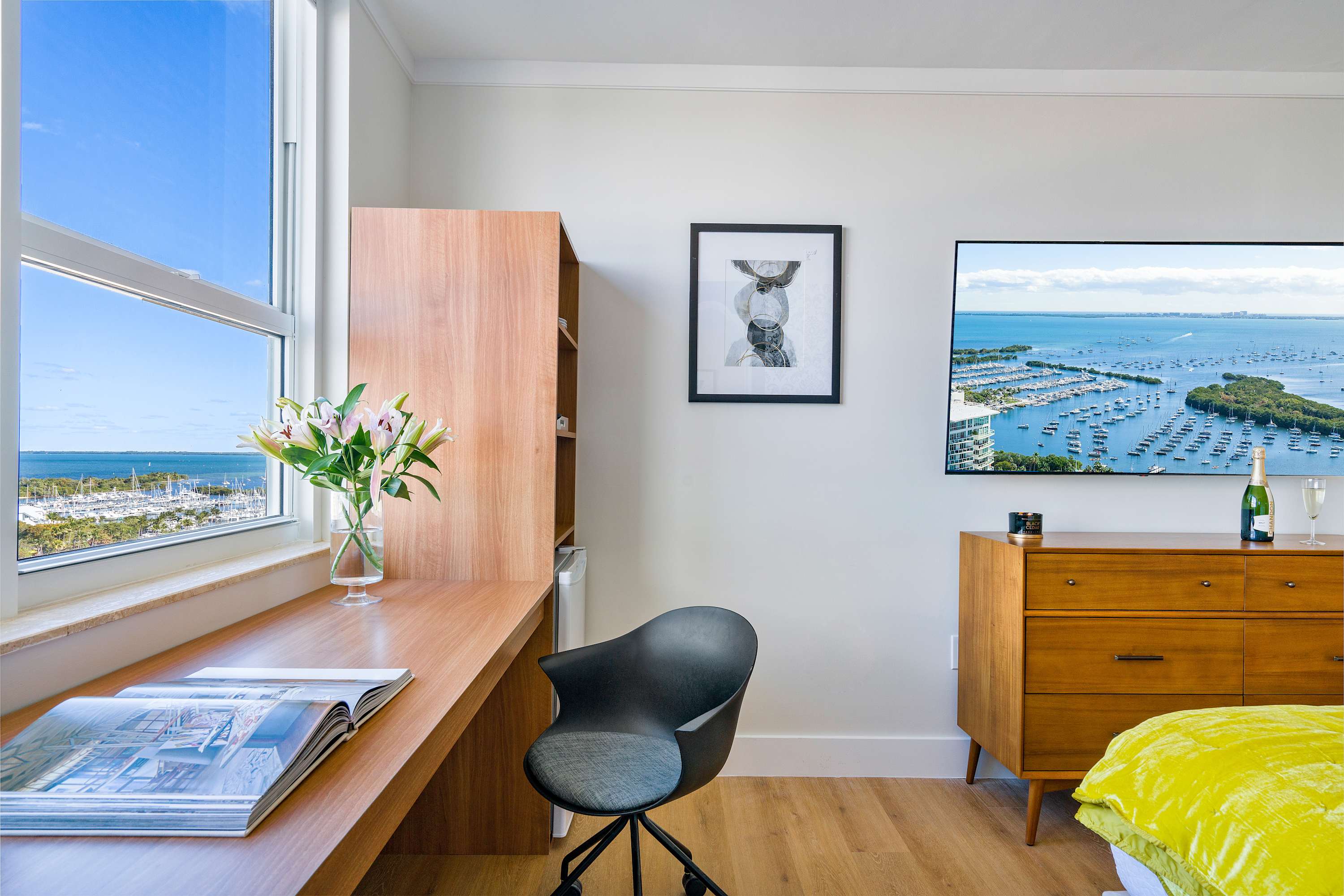 Modern bedroom corner with a long wooden desk and a black office chair positioned by a large window, showcasing a stunning bay view with boats, and a wall-mounted flat-screen TV.