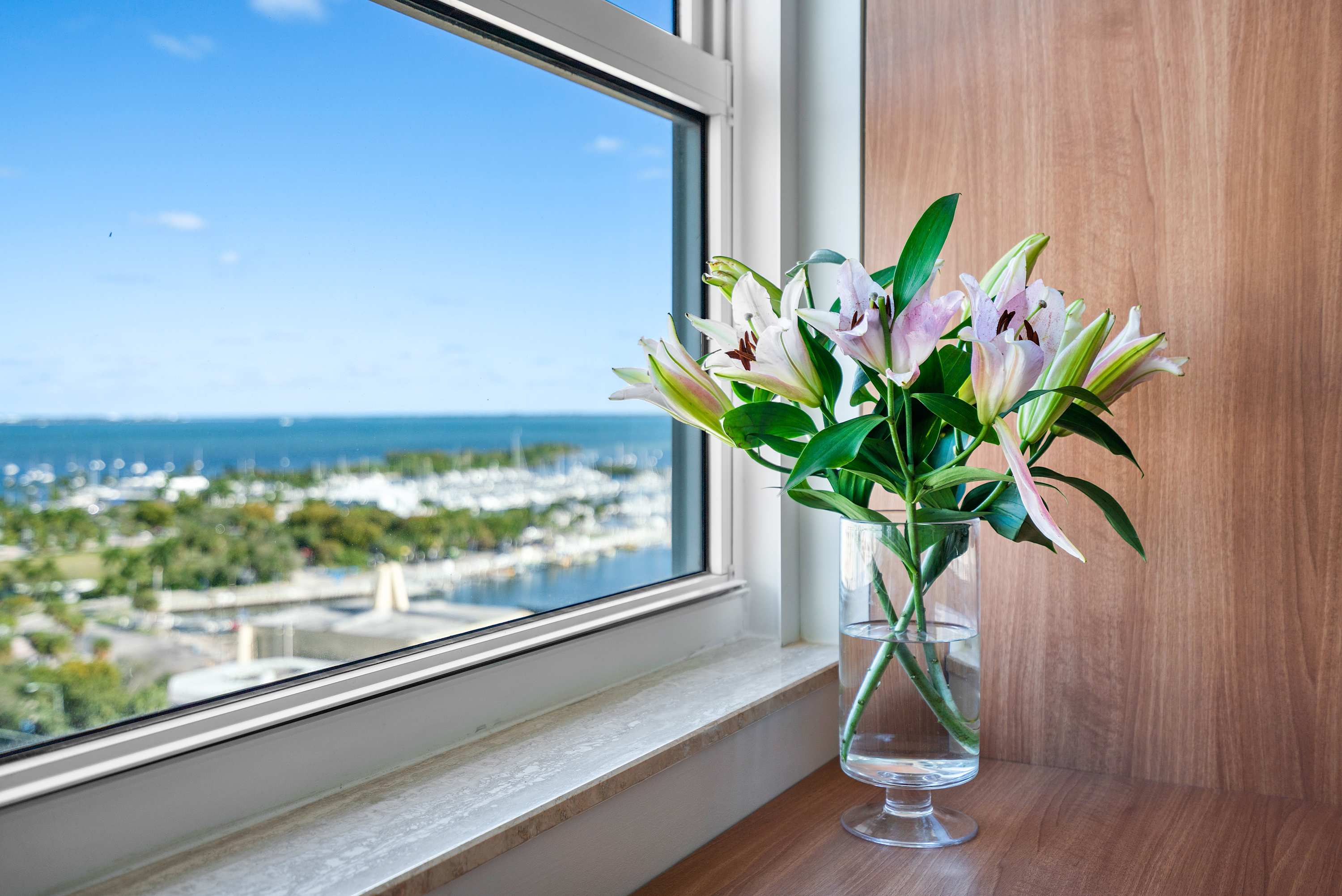 Close-up shot of a vase of fresh pink and white lilies placed on a windowsill, with a vibrant blue bay and numerous boats visible through the window in the background.