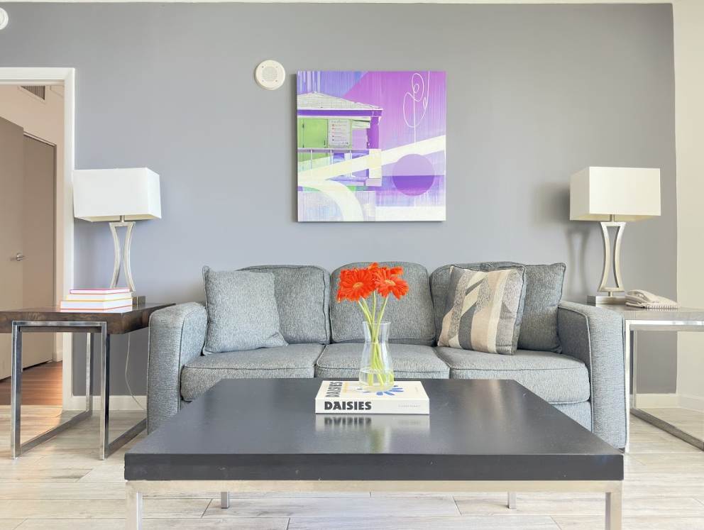 A cozy living room with a grey sofa, two end tables with modern lamps, and a black coffee table adorned with a vase of orange flowers. A contemporary abstract painting hangs on the light grey wall, complementing the light wood-look flooring.