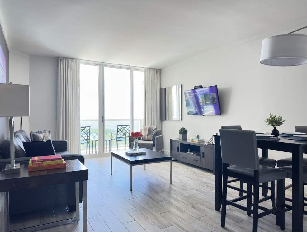 A spacious open-concept living and dining area with light wood-look flooring and floor-to-ceiling glass doors leading to a balcony. The room features a grey sofa, an armchair, a coffee table, and a high-top dining table with four chairs.