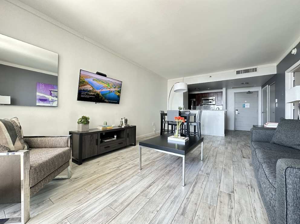 An open-concept living area with light wood-look flooring, a grey sofa, and a dark wood entertainment console with a wall-mounted TV. A high-top dining table with four chairs is visible in the background, adjacent to the modern kitchen.