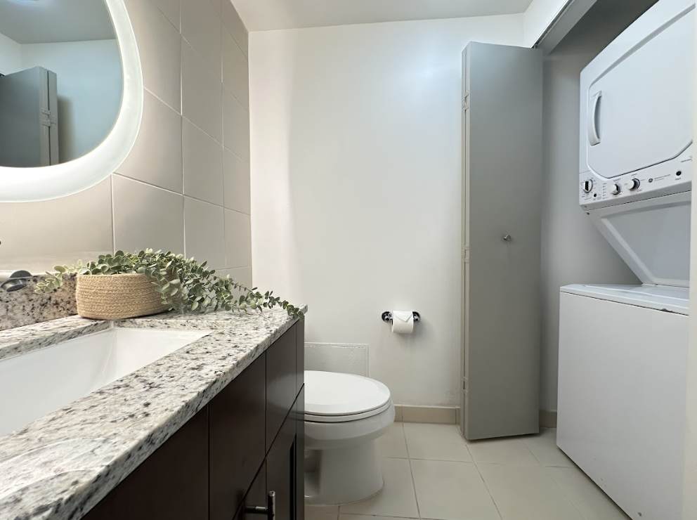 A compact bathroom featuring a single vanity with a granite countertop and a modern illuminated round mirror. The room also includes a toilet and a stacked white washer and dryer unit in a closet, with light tiled flooring.
