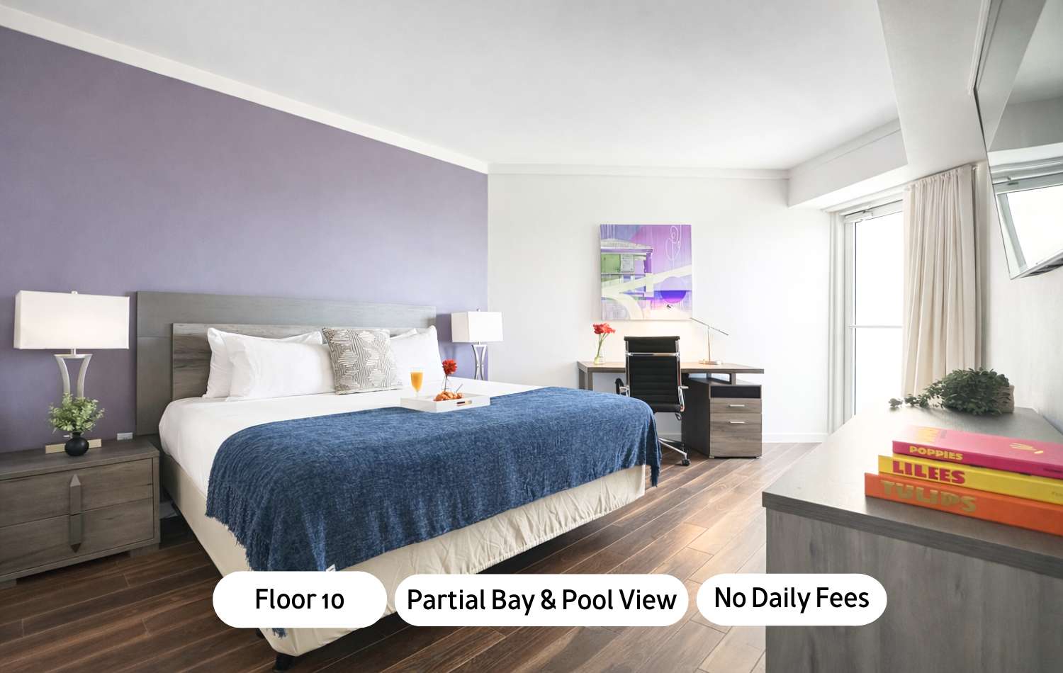 A modern bedroom featuring a king-sized bed with a grey headboard, white bedding, and a blue throw blanket. The room has light wood flooring, a purple accent wall, a desk area with a chair, and a large window with sheer curtains.