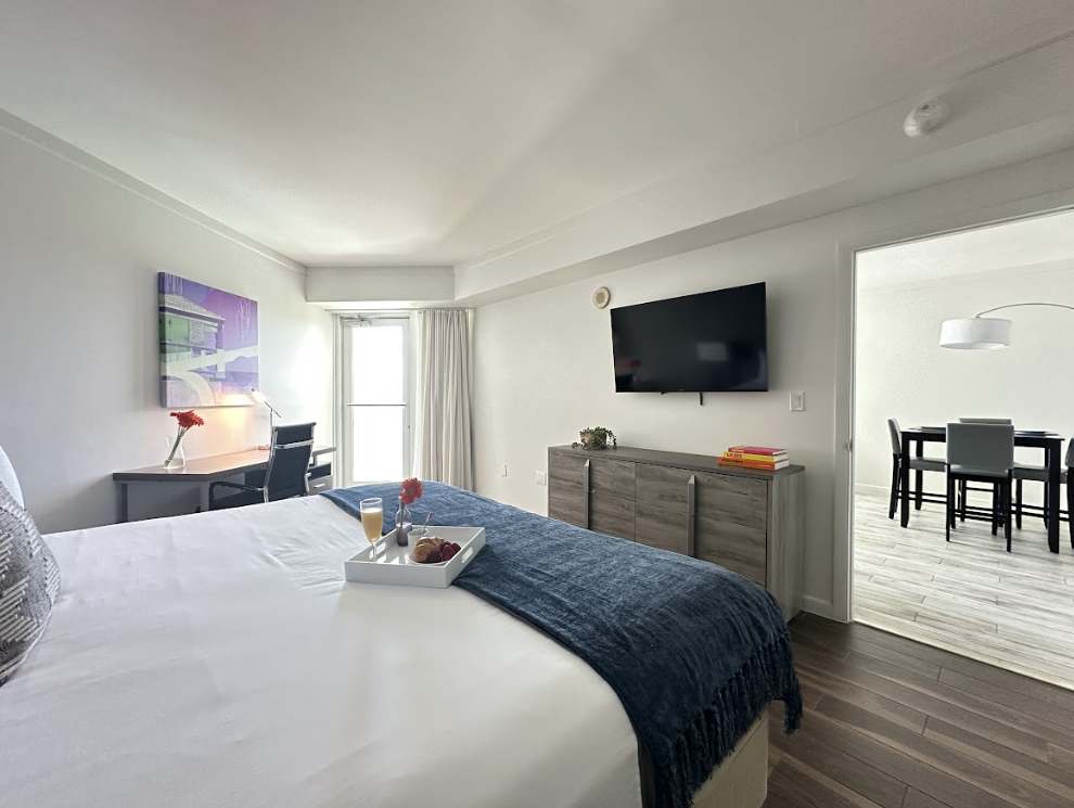 A modern bedroom featuring a king-sized bed with white bedding and a blue throw, facing a wall-mounted flat-screen TV above a grey console. The room has light wood flooring and an open doorway leading to the adjacent living and dining area.