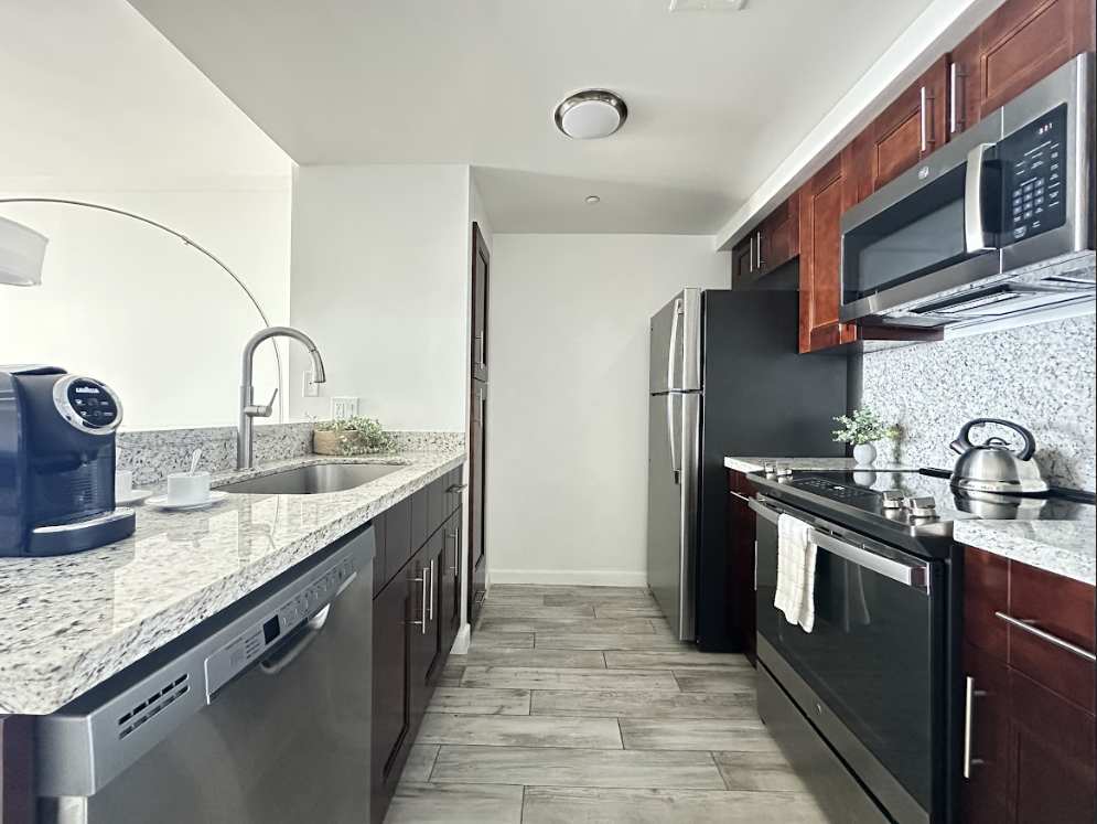 A sleek galley kitchen with dark wood cabinetry, light granite countertops, and stainless steel appliances including a dishwasher, refrigerator, and microwave. The kitchen features light wood-look flooring and a modern coffee machine on the counter.