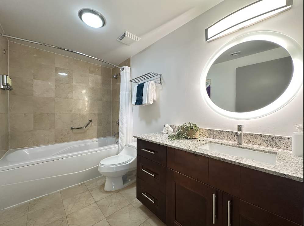 A full bathroom with a single vanity featuring a dark wood cabinet and granite countertop, a large illuminated round mirror, and a toilet. The room also has a bathtub with a shower, tiled walls, and light tiled flooring.
