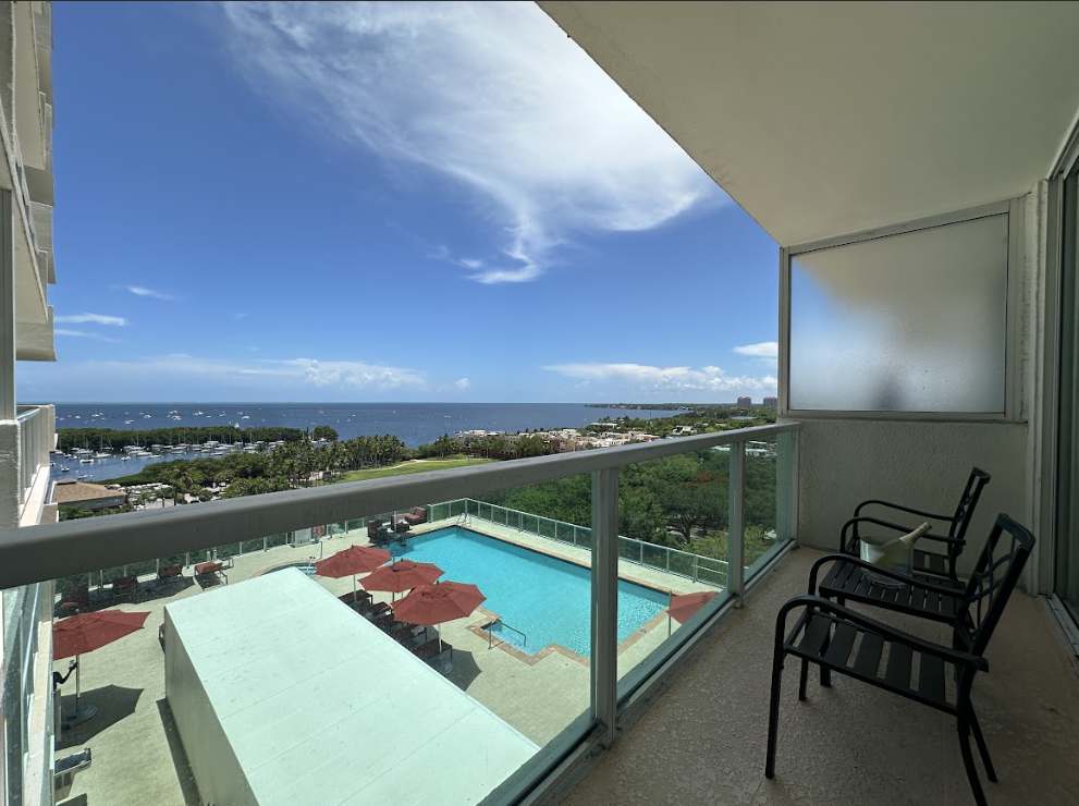 A private balcony with clear glass railings, offering panoramic views of a sparkling swimming pool, lush green landscape, and a vast bay dotted with boats. Two black outdoor chairs provide a comfortable spot to enjoy the scenic vista.