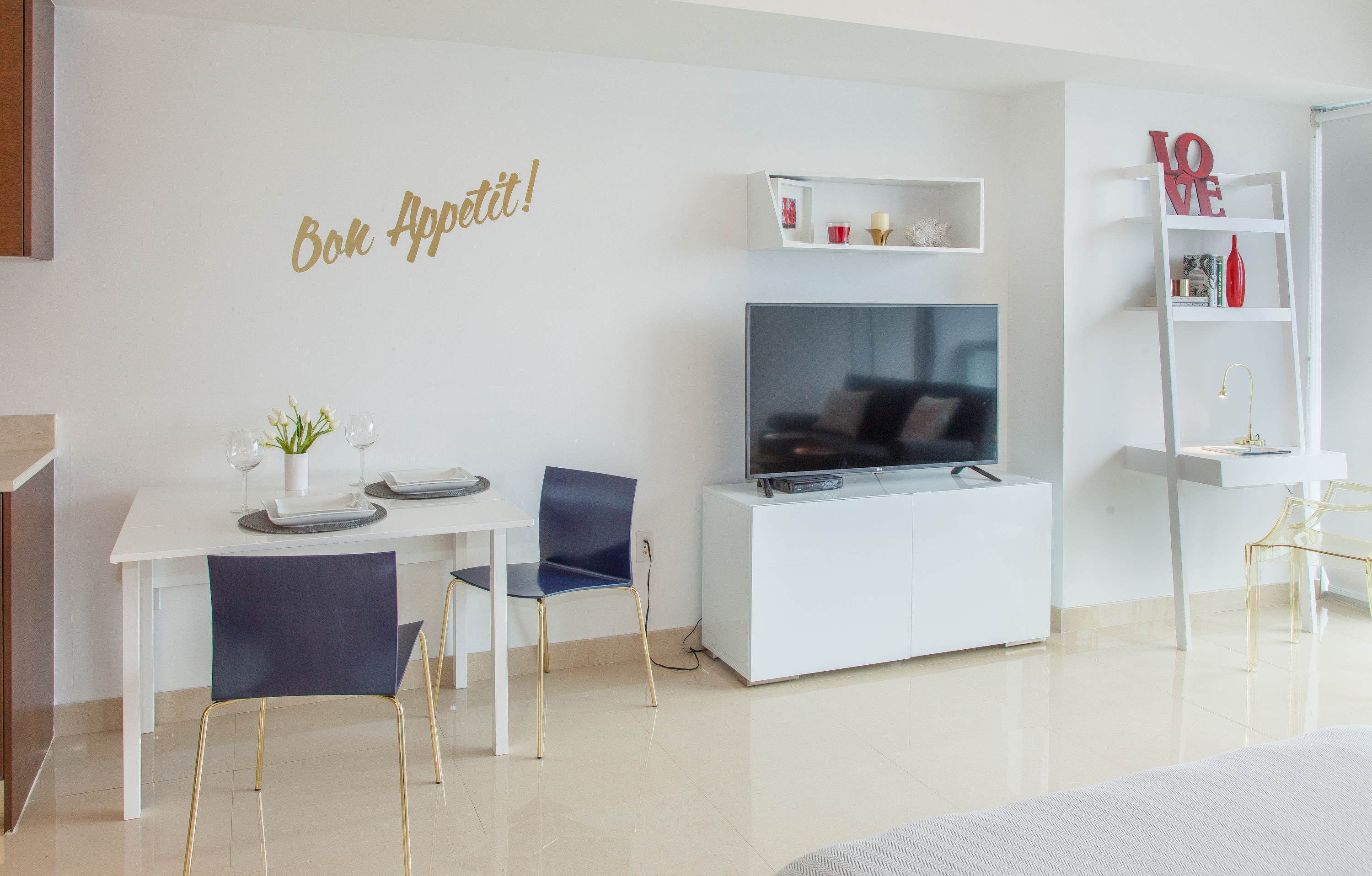 A bright dining area with a white rectangular table and two dark chairs, adjacent to a living space featuring a flat-screen TV on a white console and decorative wall shelves. A "Bon Appetit!" decal adorns the wall.