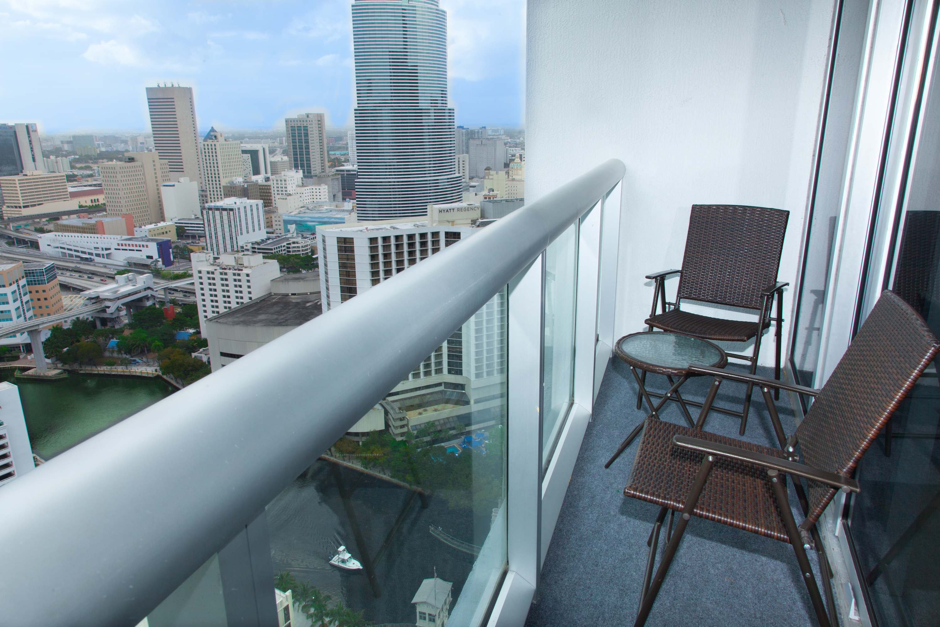 A private balcony furnished with two wicker chairs and a small glass-top table, offering breathtaking panoramic views of a city skyline and a river with boats. The railing is glass for unobstructed sightlines.