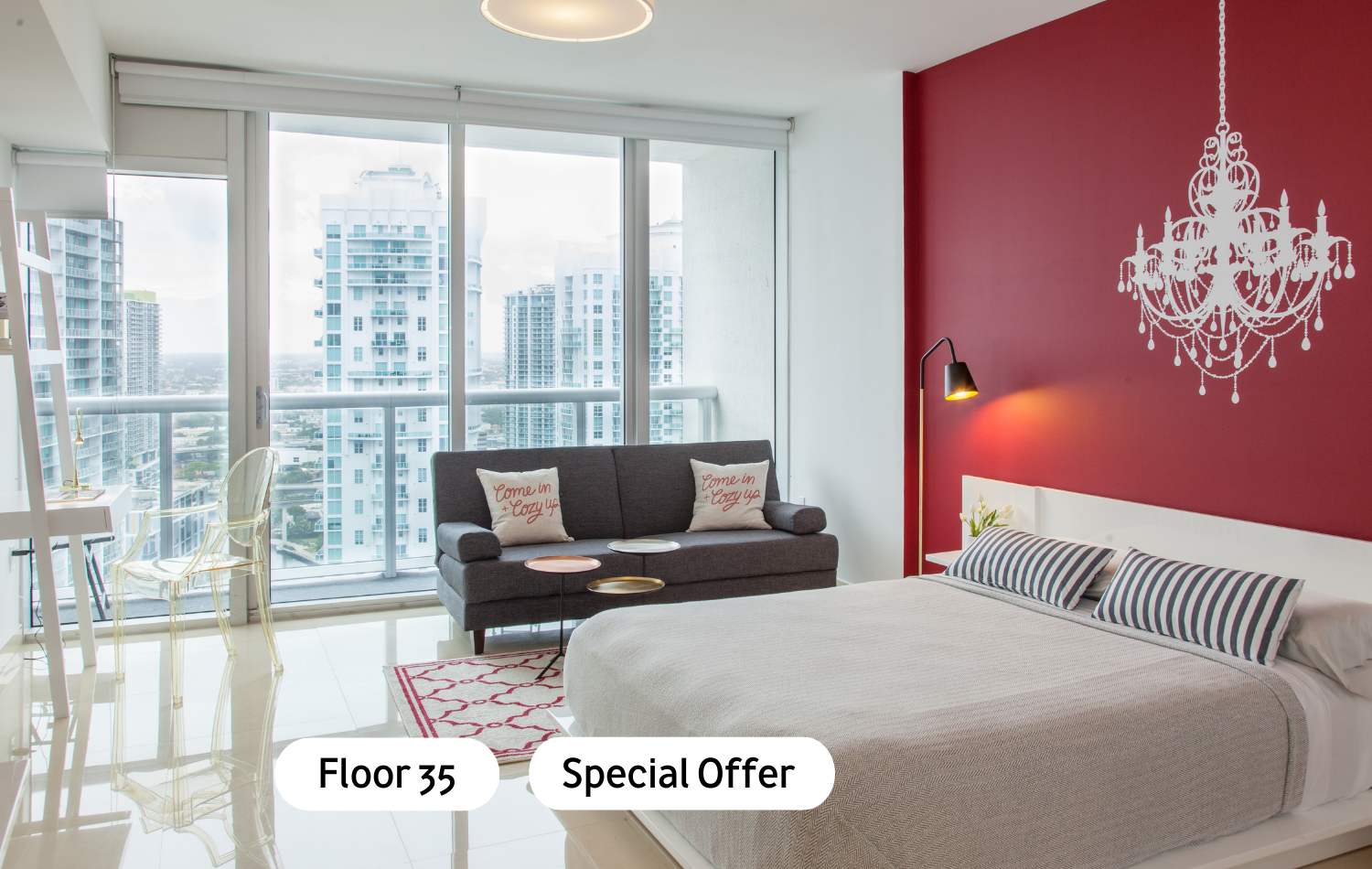 A modern studio apartment featuring a queen-sized bed with striped pillows, a dark grey sofa, and a white desk, all set against a red accent wall with a white chandelier decal. Floor-to-ceiling windows offer expansive city views.