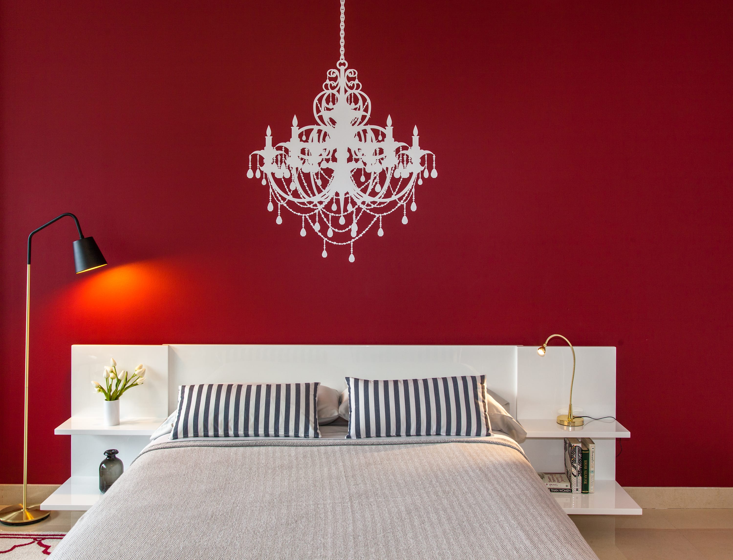Close-up of a modern bedroom area with a queen bed, striped pillows, and white nightstands, set against a striking red accent wall adorned with a large white chandelier decal. Two stylish lamps provide ambient lighting.