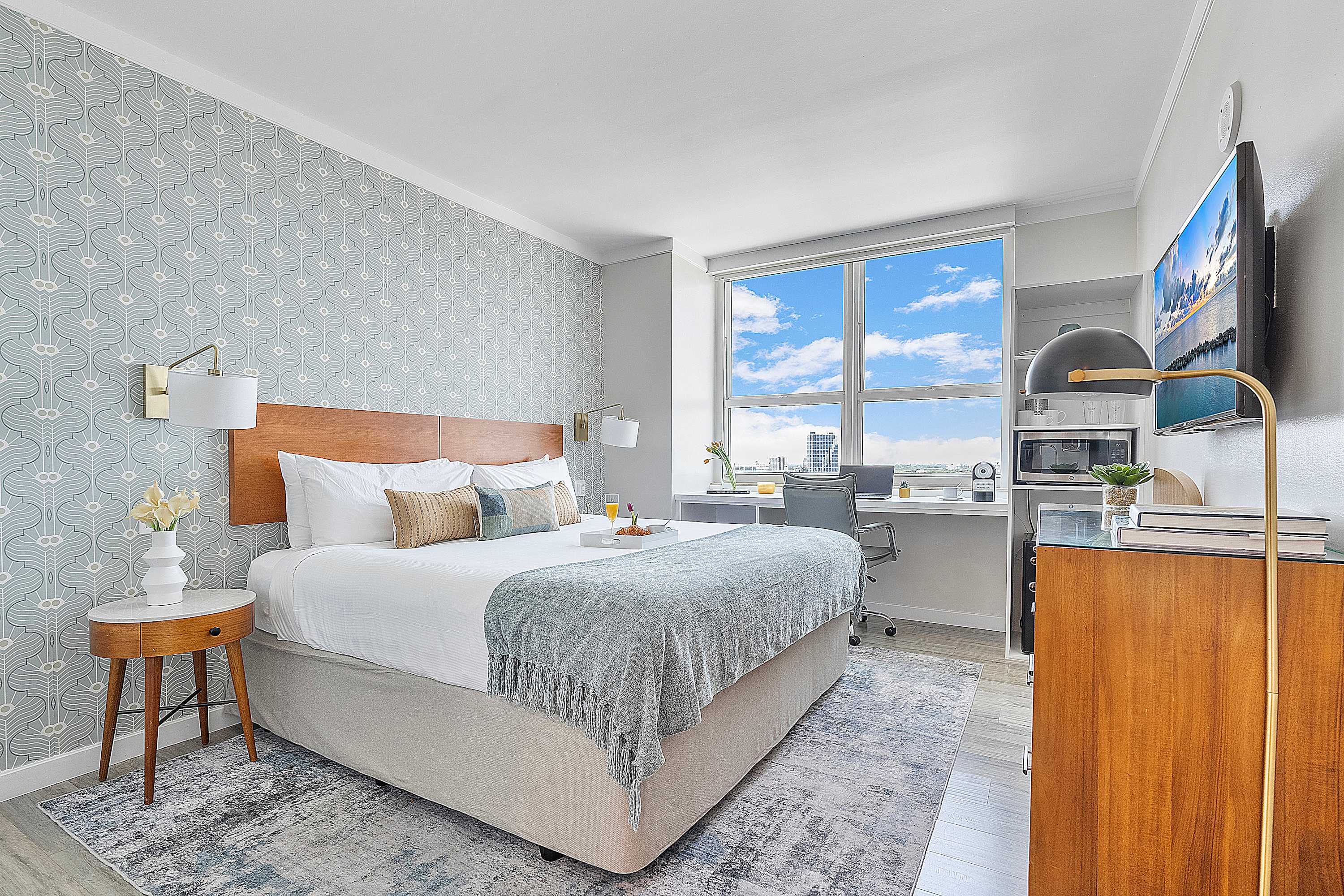 Cozy bedroom-2 featuring a queen-sized bed with a wooden headboard and light blue bedding, an area rug, and a large window offering city views. A built-in desk with a chair is positioned by the window, and a wall-mounted TV is visible.