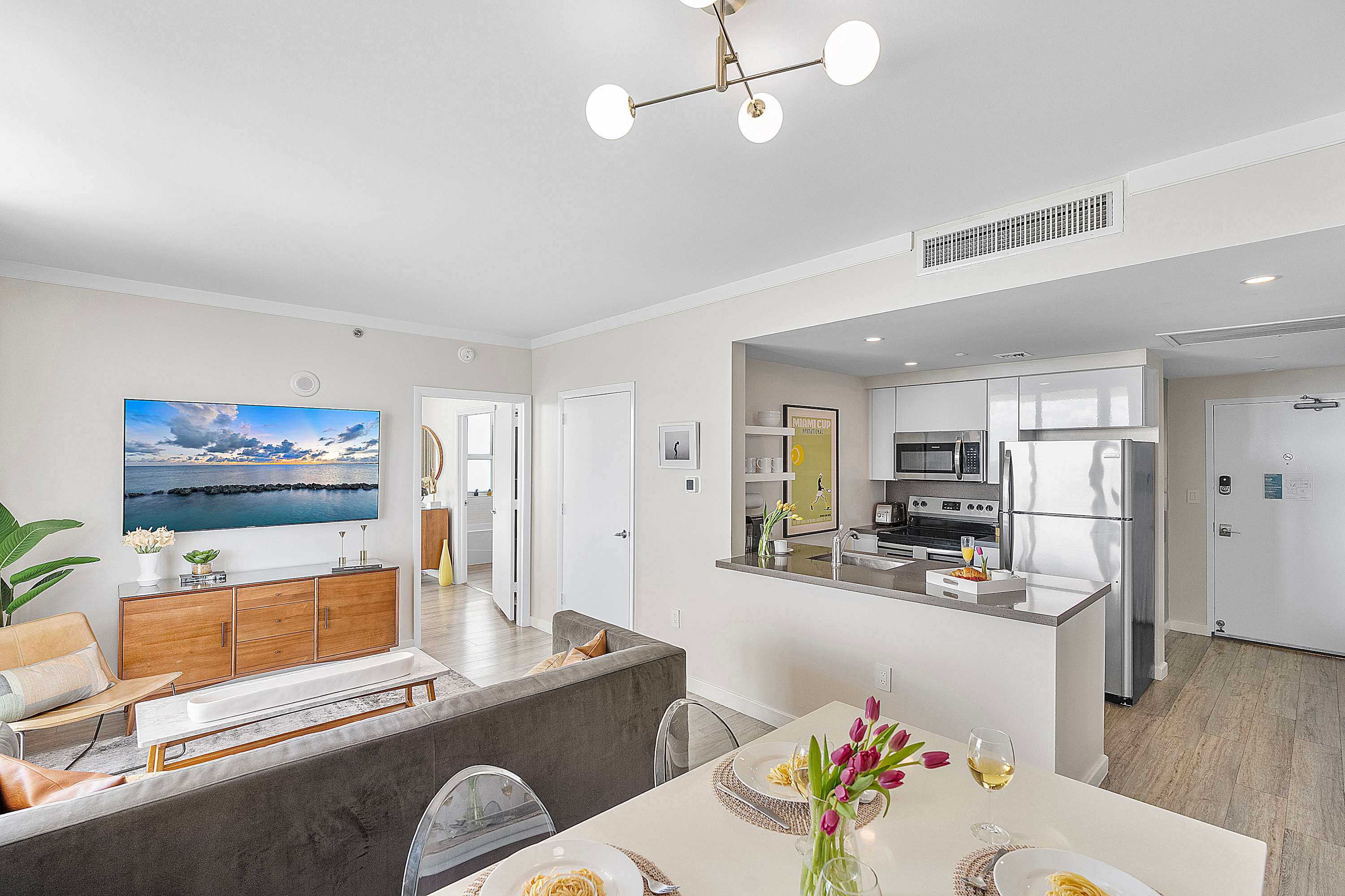 Bright open-plan living space featuring a gray sofa, a wall-mounted flat-screen TV above a wooden console, and a dining table with clear chairs, with a modern kitchen visible in the background.