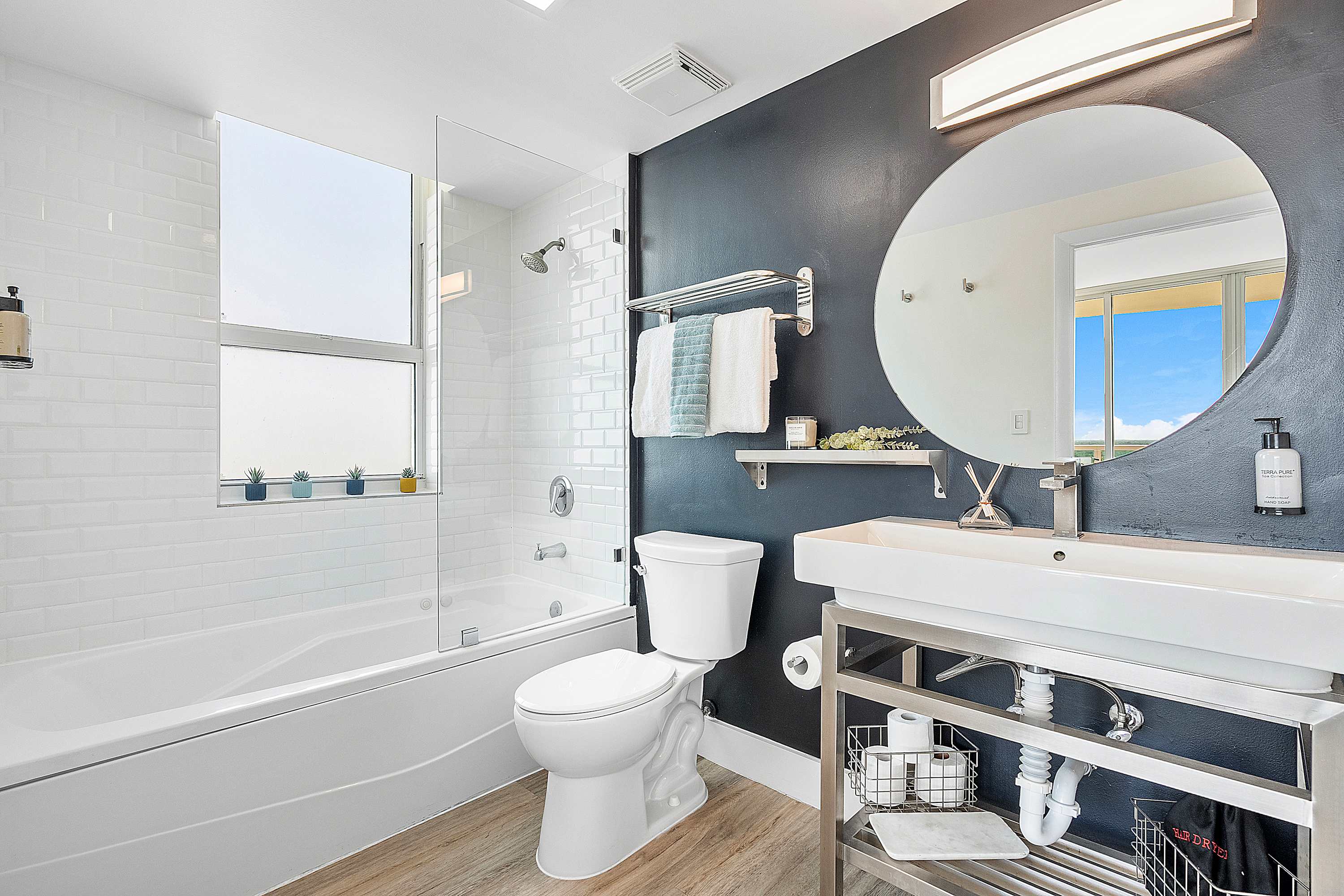 Modern bathroom-1 featuring a white subway-tiled shower/tub combo with a glass partition, a dark accent wall behind a round mirror, and a contemporary vanity with an open metal frame. A window provides natural light.