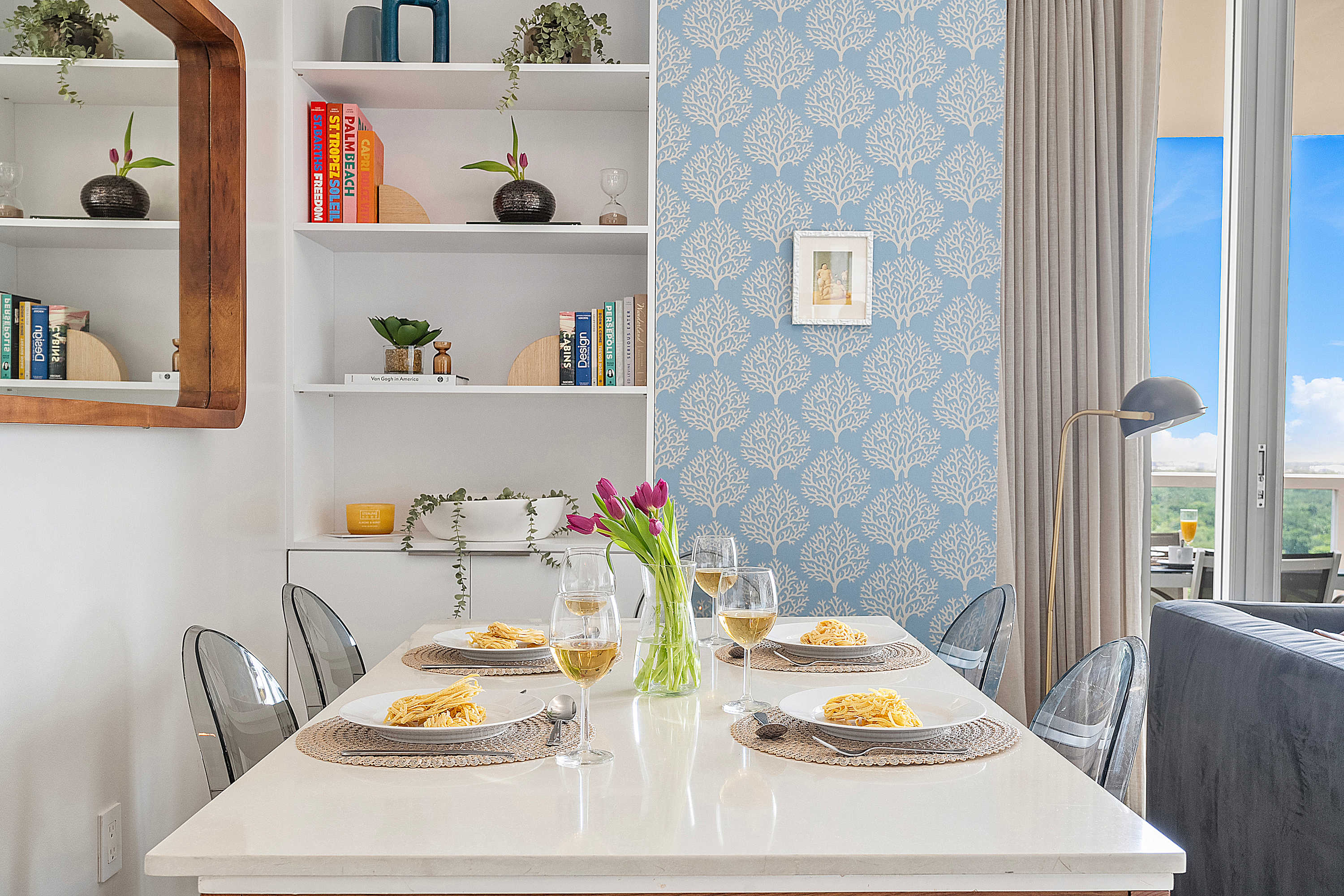 Elegant dining area featuring a white rectangular table set for four with clear acrylic chairs, against a backdrop of white built-in shelves and a light blue patterned accent wall. A vase of purple tulips adds a pop of color.