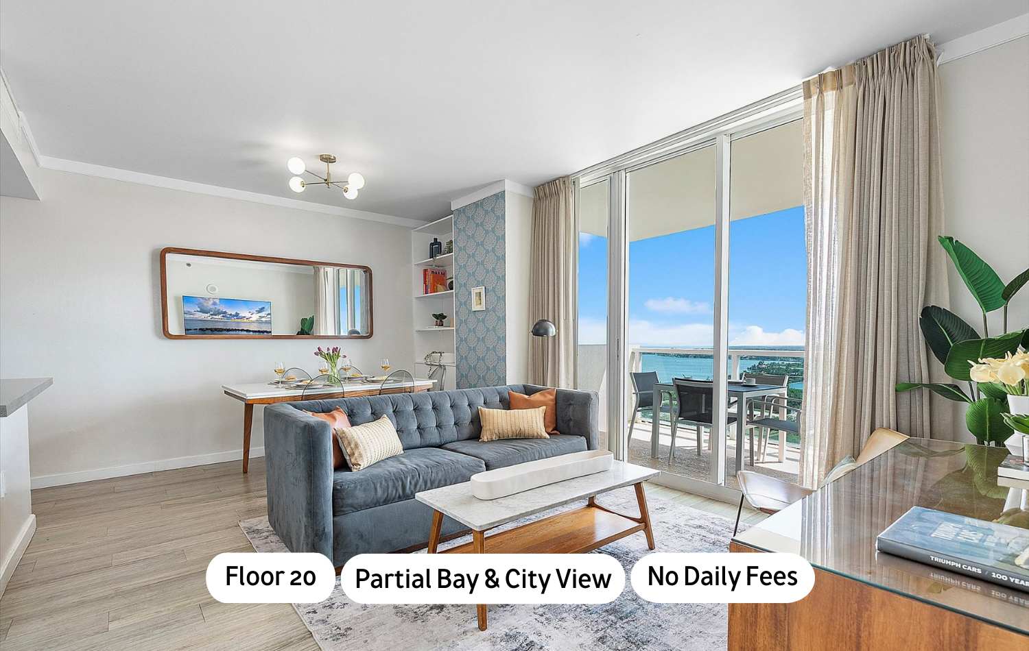 Modern open-concept living and dining area featuring a gray tufted sofa, a white marble coffee table, and a dining table with clear chairs, all illuminated by a contemporary chandelier. Large sliding glass doors open to a balcony with ocean and city views.