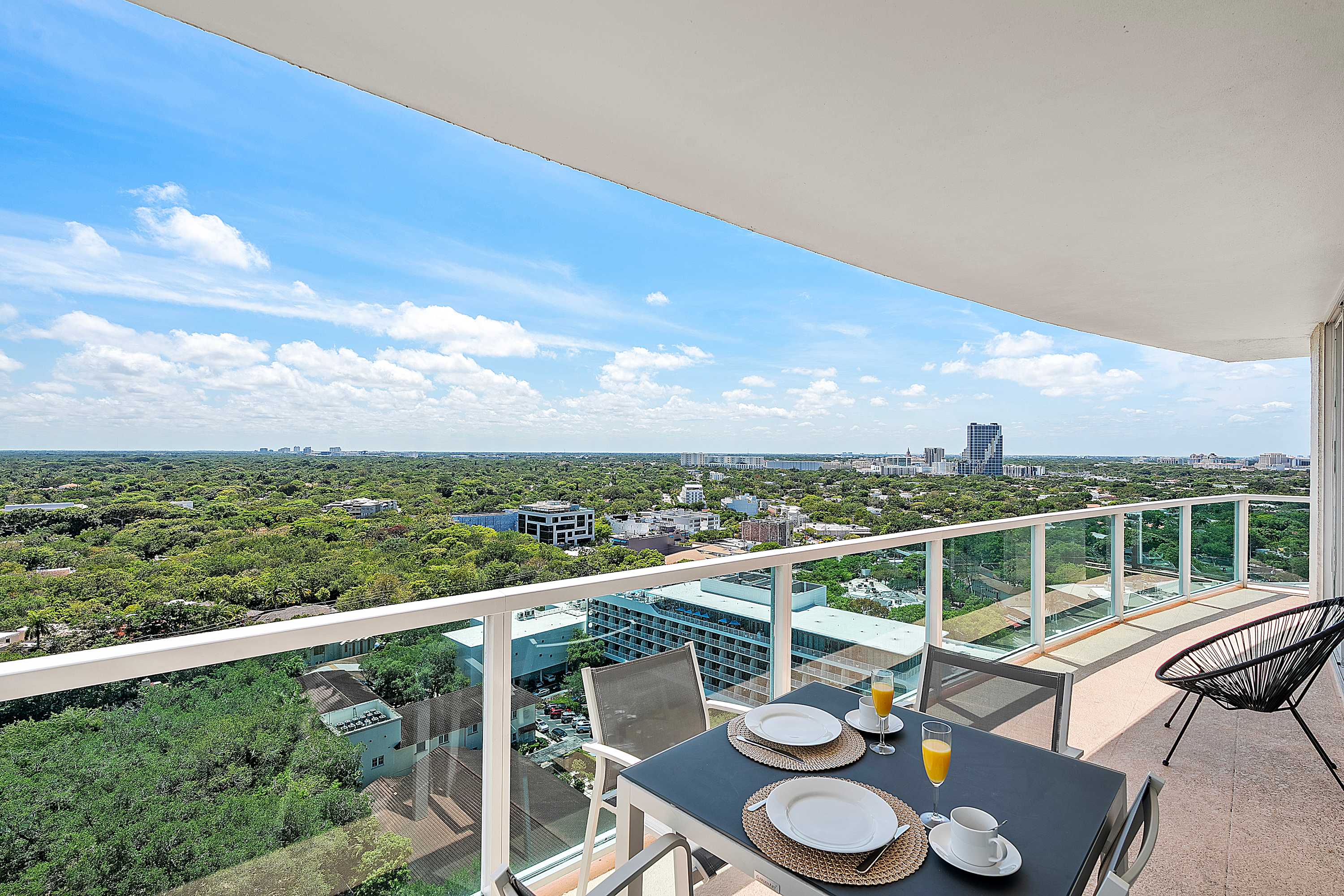 Spacious balcony with a small outdoor dining set, offering expansive panoramic views of a lush green city, distant buildings, and a wide body of water under a bright blue sky.
