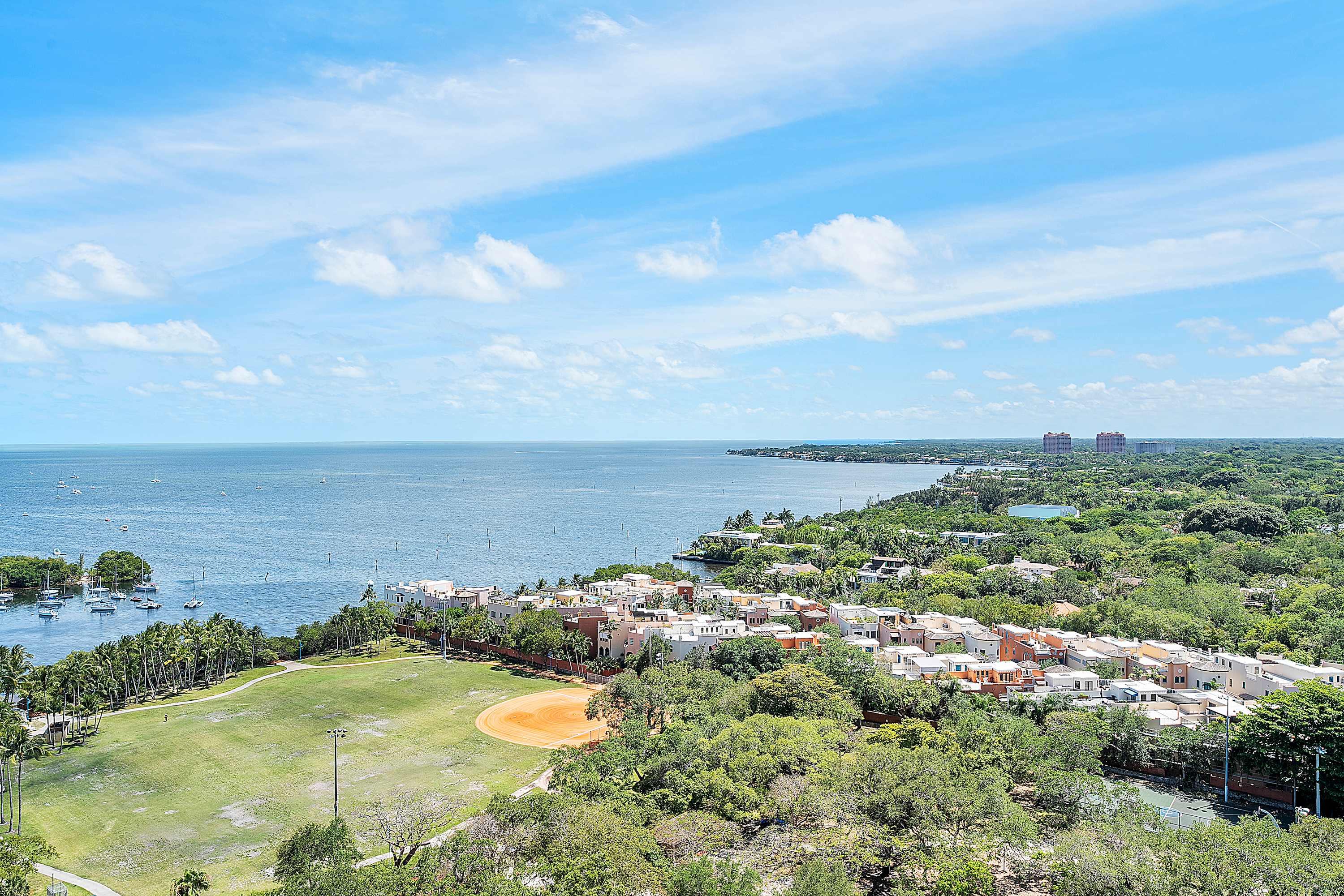 Expansive aerial view showcasing a vibrant blue bay dotted with boats, a lush green baseball field, and a residential area with colorful buildings under a bright blue sky with scattered clouds. The coastline stretches into the distance with more urban development.