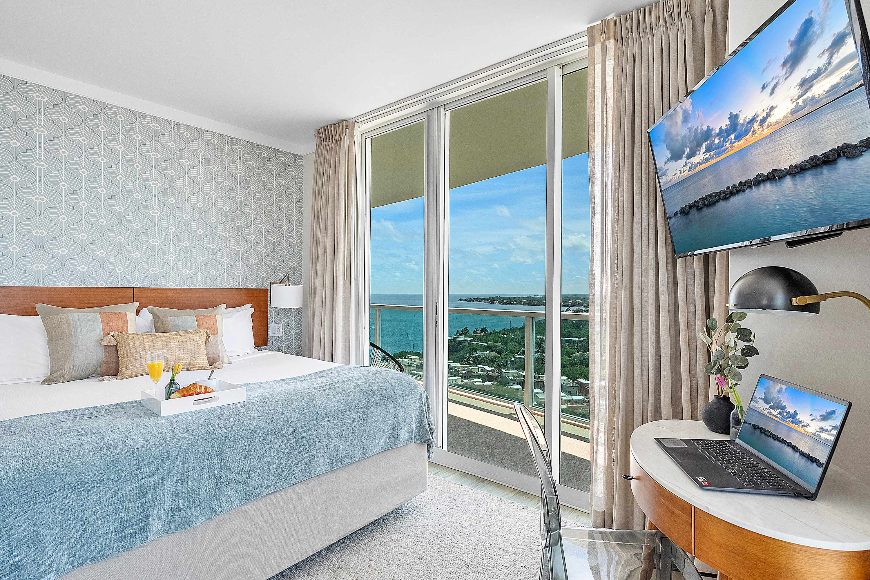 Bright bedroom-1 with a queen-sized bed, light blue bedding, and a large sliding glass door leading to a private balcony with expansive ocean and city views. A wall-mounted TV and a small desk area are also visible.