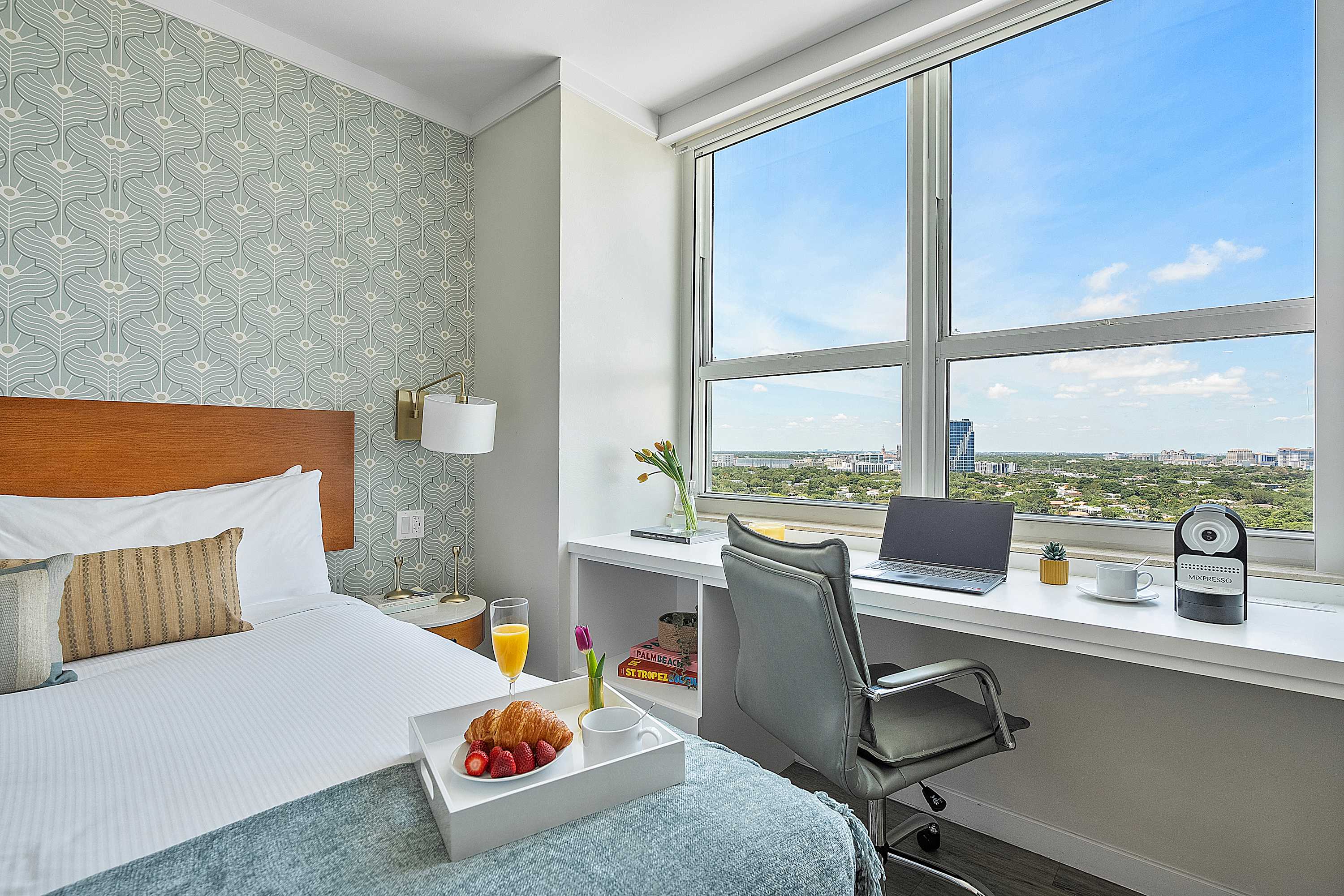 Detailed view of bedroom-2's workspace, featuring a built-in white desk with a laptop and coffee machine, positioned by a large window offering expansive city views. The queen bed with light blue bedding and a patterned accent wall are also visible.