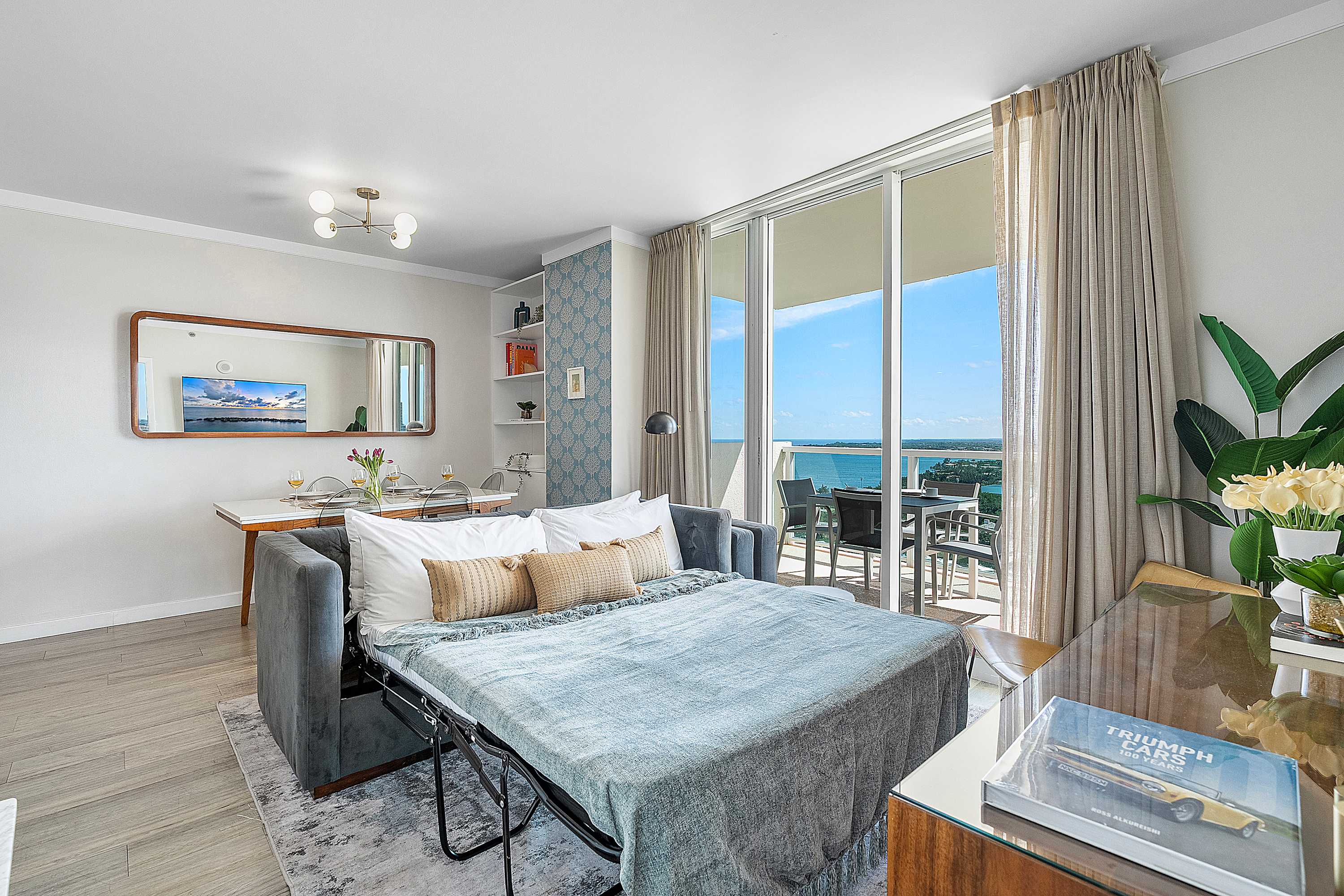 Living room with a gray sofa converted into a comfortable sofa bed, facing a large sliding glass door that opens to a balcony with a stunning ocean view. A dining area is visible in the background.