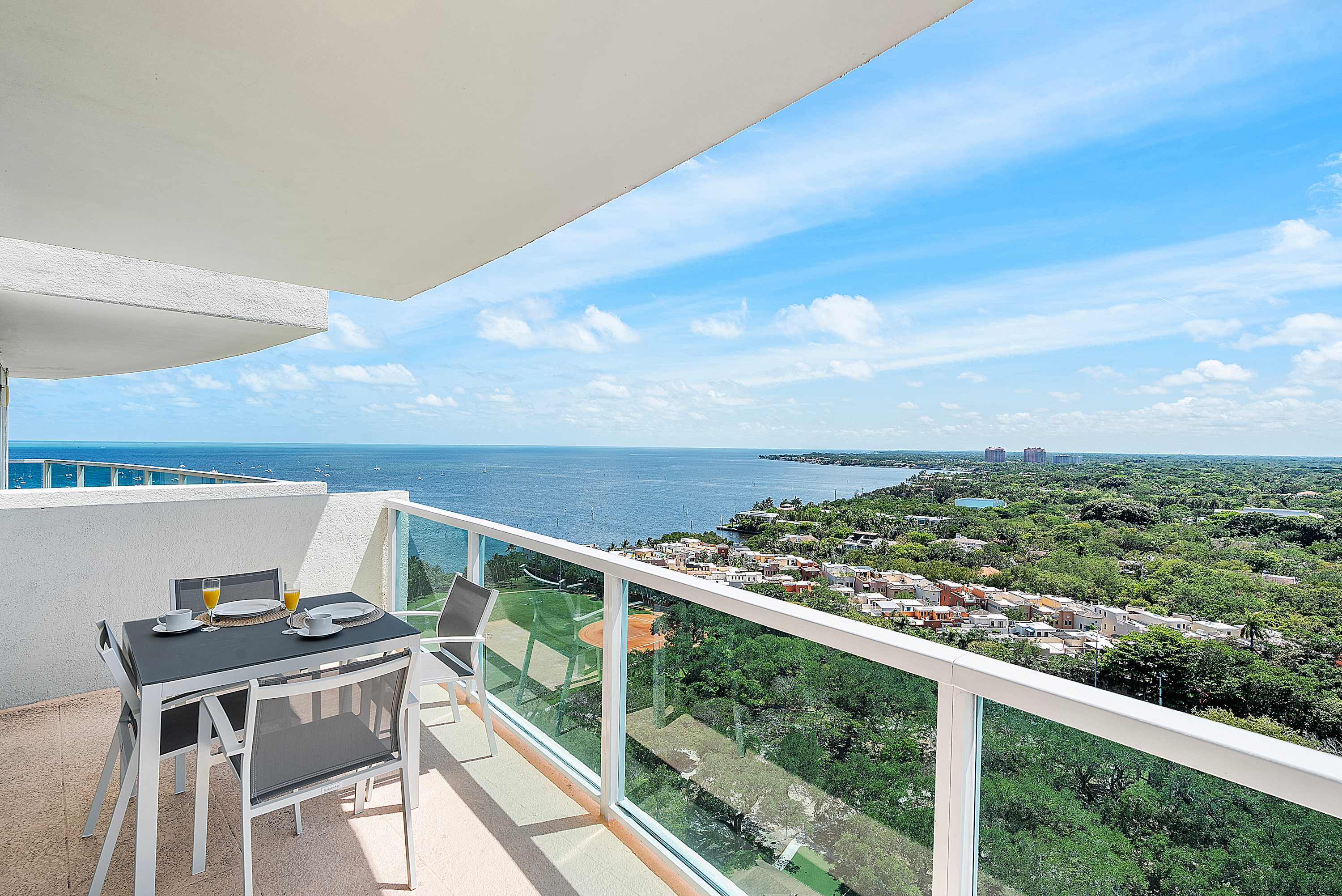 Expansive balcony with an outdoor dining set, offering breathtaking panoramic views of the vast ocean meeting the horizon, with a glimpse of the city and lush coastline below.