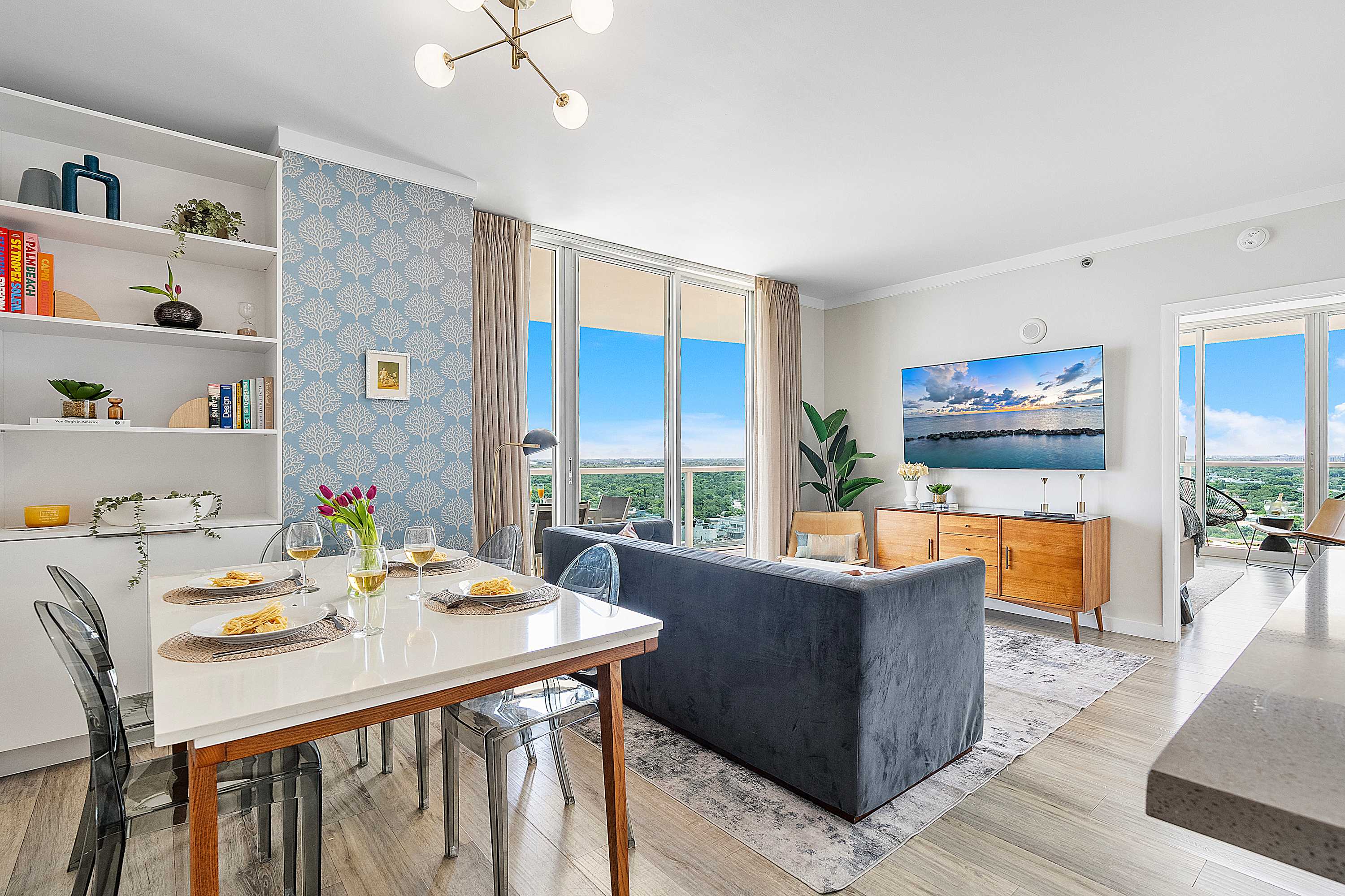 Modern open-concept living and dining area featuring a gray velvet sofa, a white dining table with clear chairs, and large sliding glass doors opening to a balcony with city and ocean views. A light blue patterned accent wall adds a pop of color.