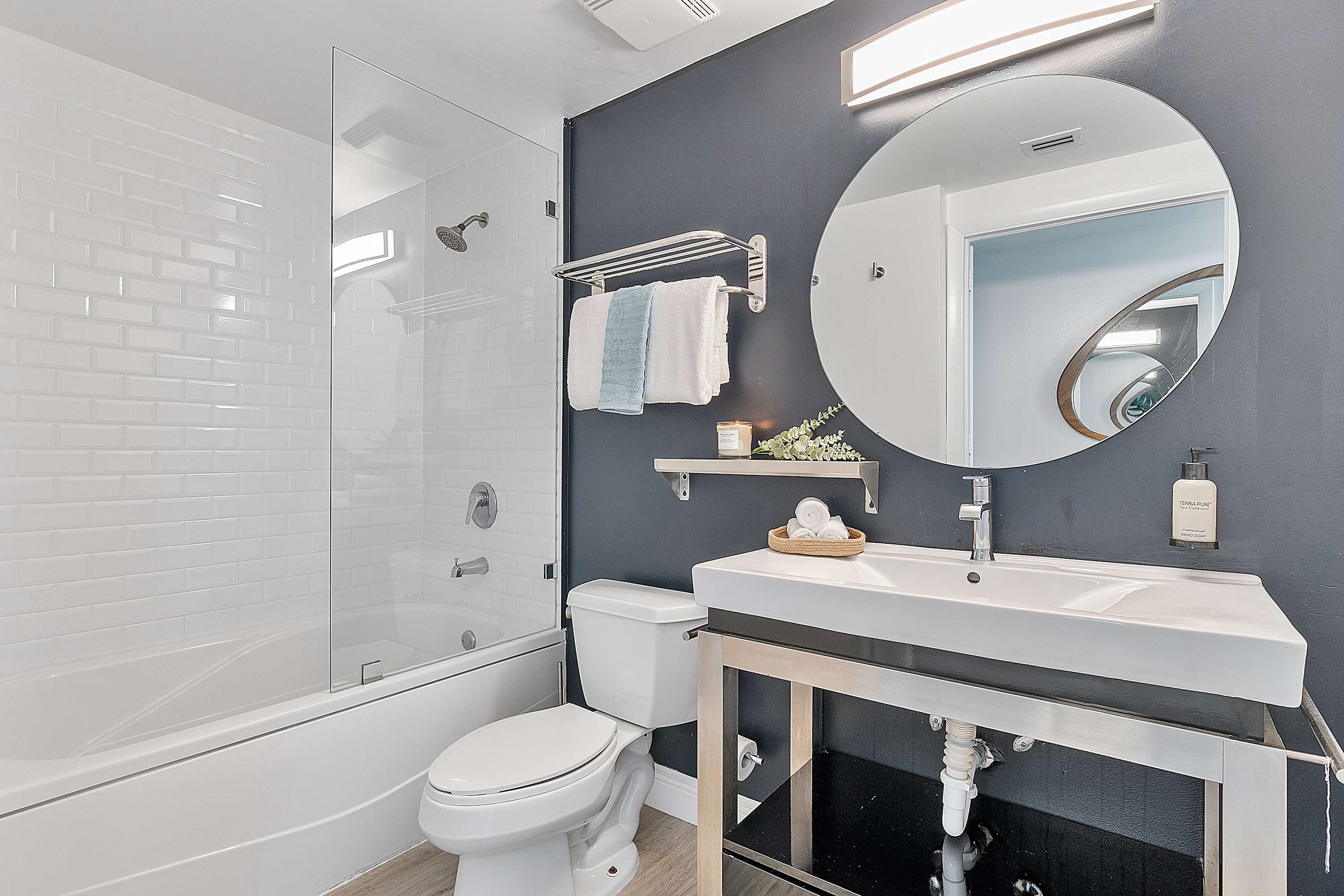 Modern bathroom-2 featuring a white subway-tiled shower/tub combo with a glass partition, a dark accent wall behind a round mirror, and a contemporary vanity with an open metal frame. The layout is clean and functional.