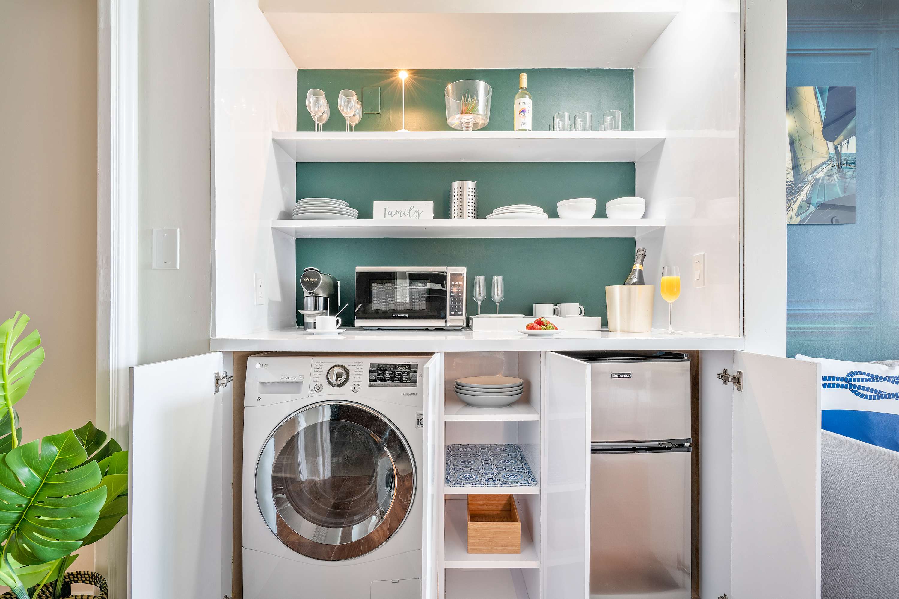 A compact kitchenette area featuring a stacked washer and dryer, a microwave, a mini-fridge, and open shelving stocked with dishes and glassware, all neatly integrated into white cabinetry.