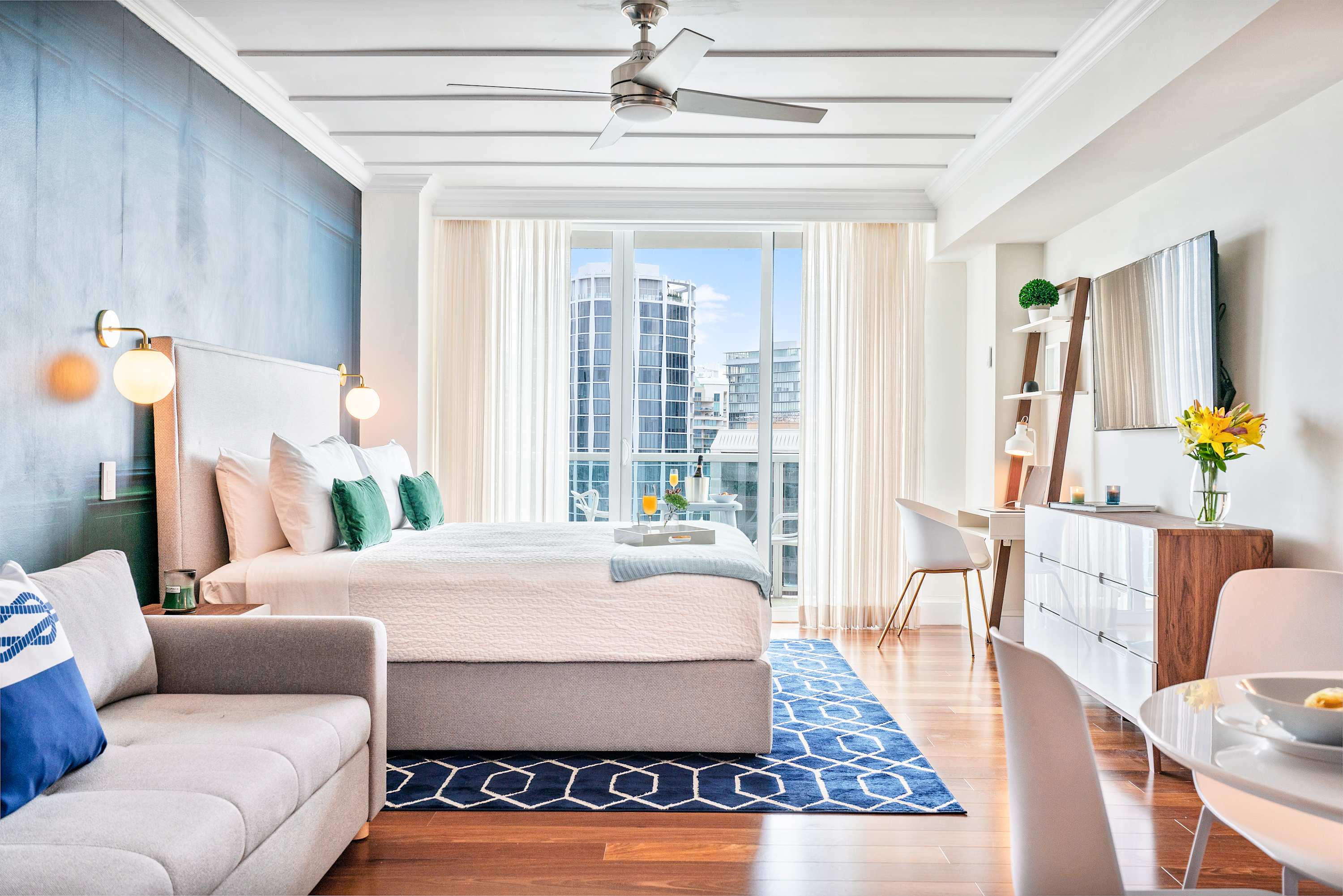 A bright studio space featuring a king bed, a small desk with a chair, and a dining table, all on polished hardwood floors with a large window offering city views.