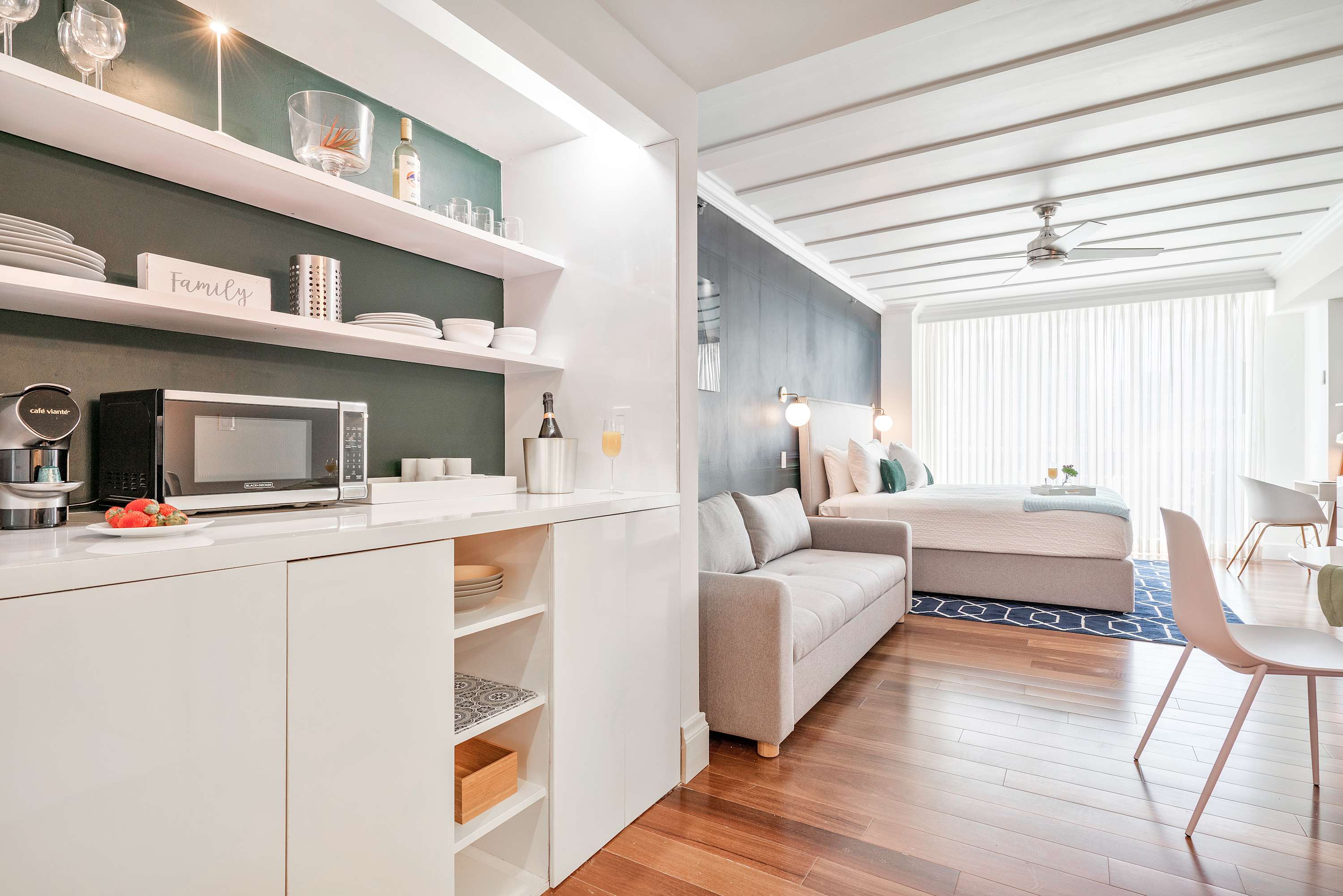 A modern kitchenette with white cabinetry, open shelves, a microwave, and a coffee maker, seamlessly integrated into the studio space with hardwood floors and a view towards the sleeping area.