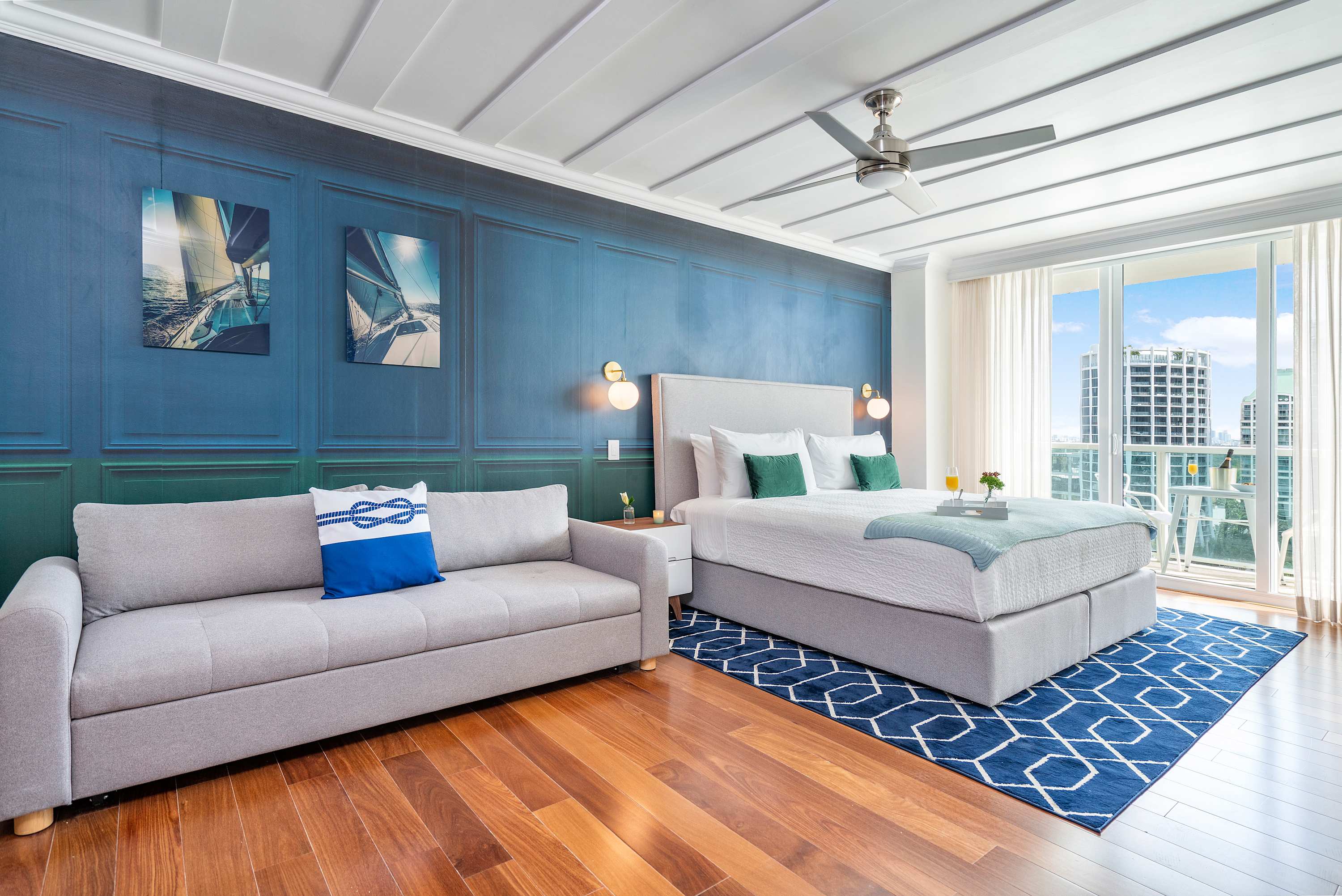 A spacious studio room with a king-sized bed on a blue geometric rug, a grey sofa, and large glass doors leading to a balcony, all under a white coffered ceiling with a fan.