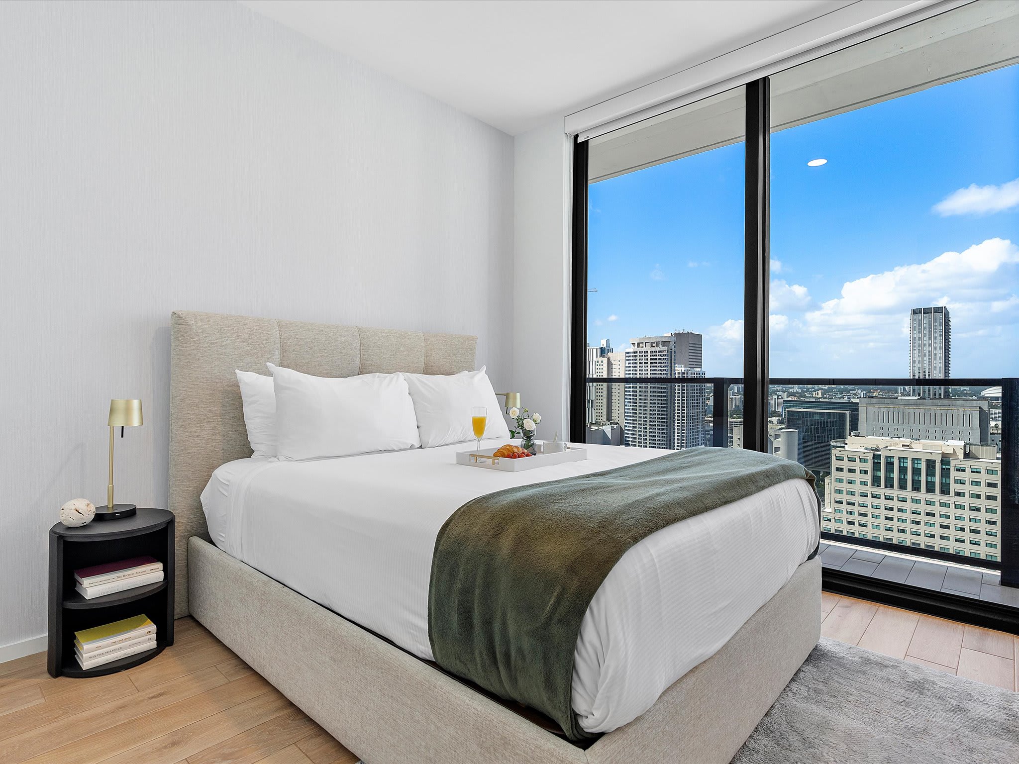 Bright bedroom with a queen-sized bed, light upholstered headboard, white bedding with a green throw, and a black round nightstand, offering city views through large windows.