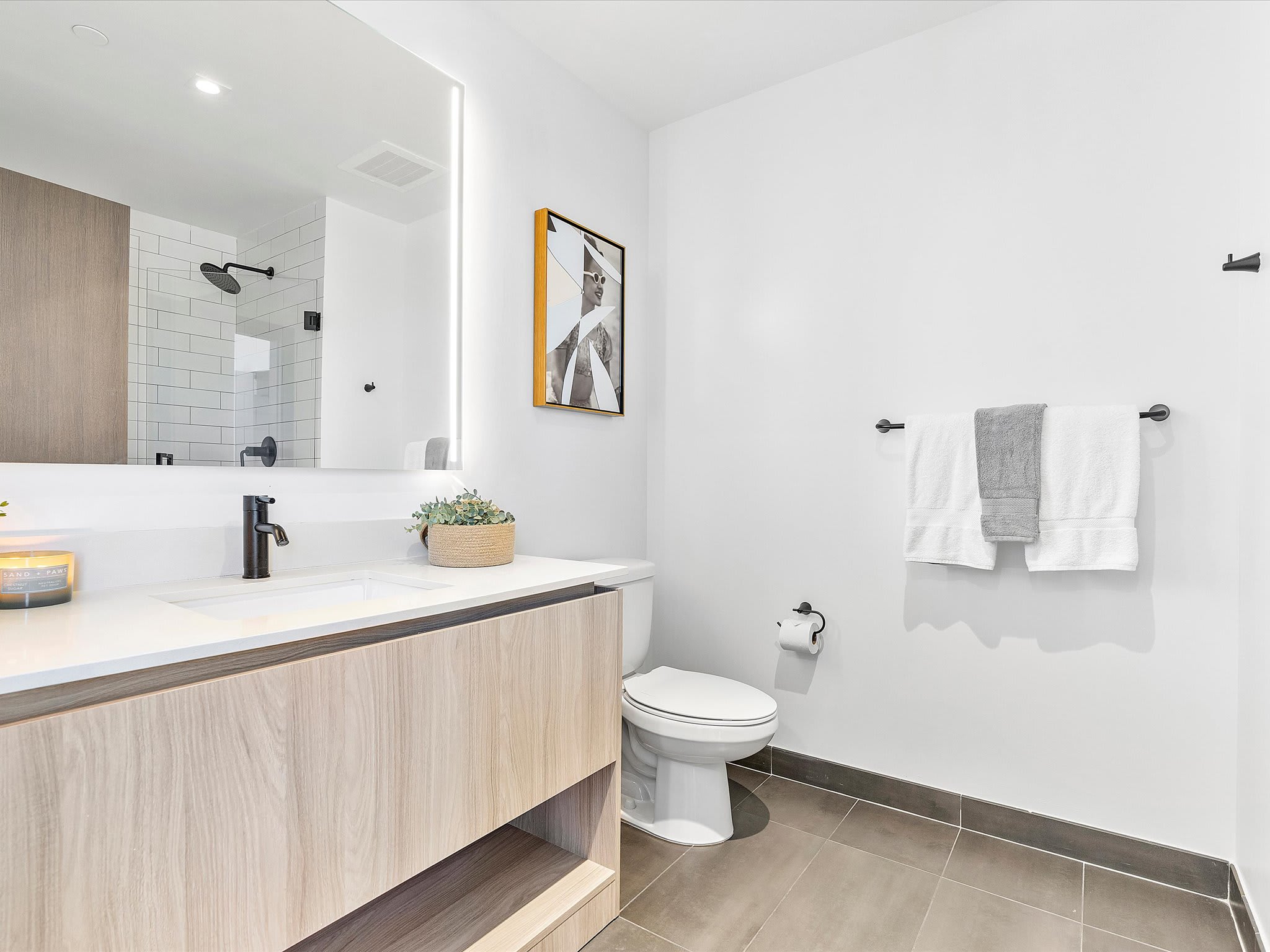 Contemporary bathroom with a light wood vanity, white countertop, black faucet, illuminated mirror, and a toilet, featuring white subway tile in the shower area and dark grey floor tiles.
