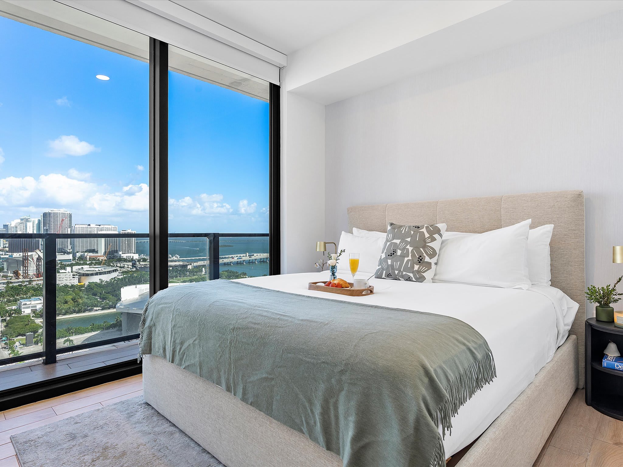 Spacious bedroom with a king-sized bed featuring a light upholstered headboard, light grey throw, and a breakfast tray, offering panoramic city and bay views through large floor-to-ceiling windows.