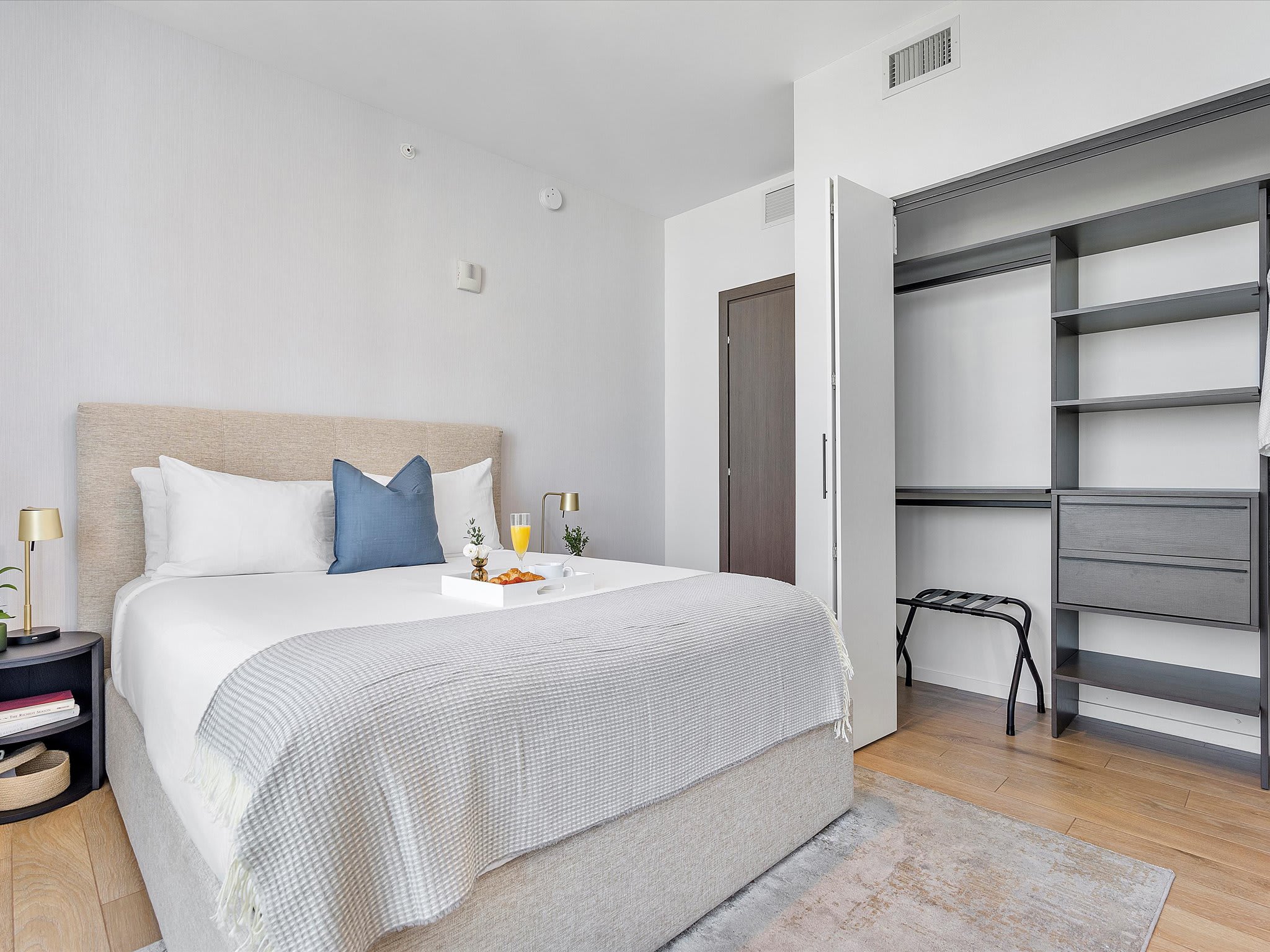Cozy bedroom with a queen-sized bed, light upholstered headboard, white bedding with a blue accent pillow, and a built-in open closet system with shelves and drawers, set on light wood flooring.