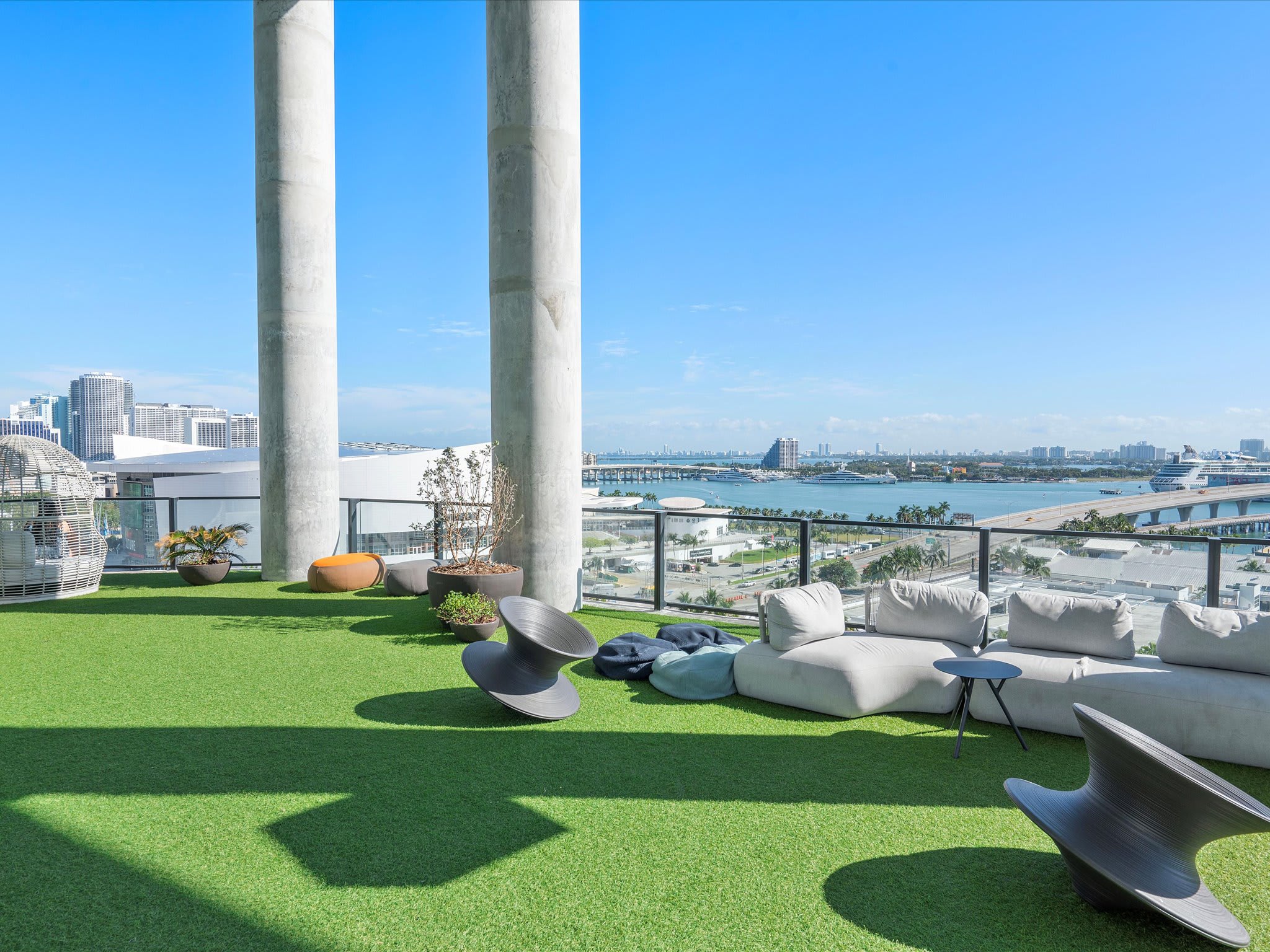 Another view of the rooftop lounge shows modern, low-profile outdoor seating on artificial turf, with large concrete pillars framing the expansive city and bay views. The clear blue sky enhances the bright outdoor setting.