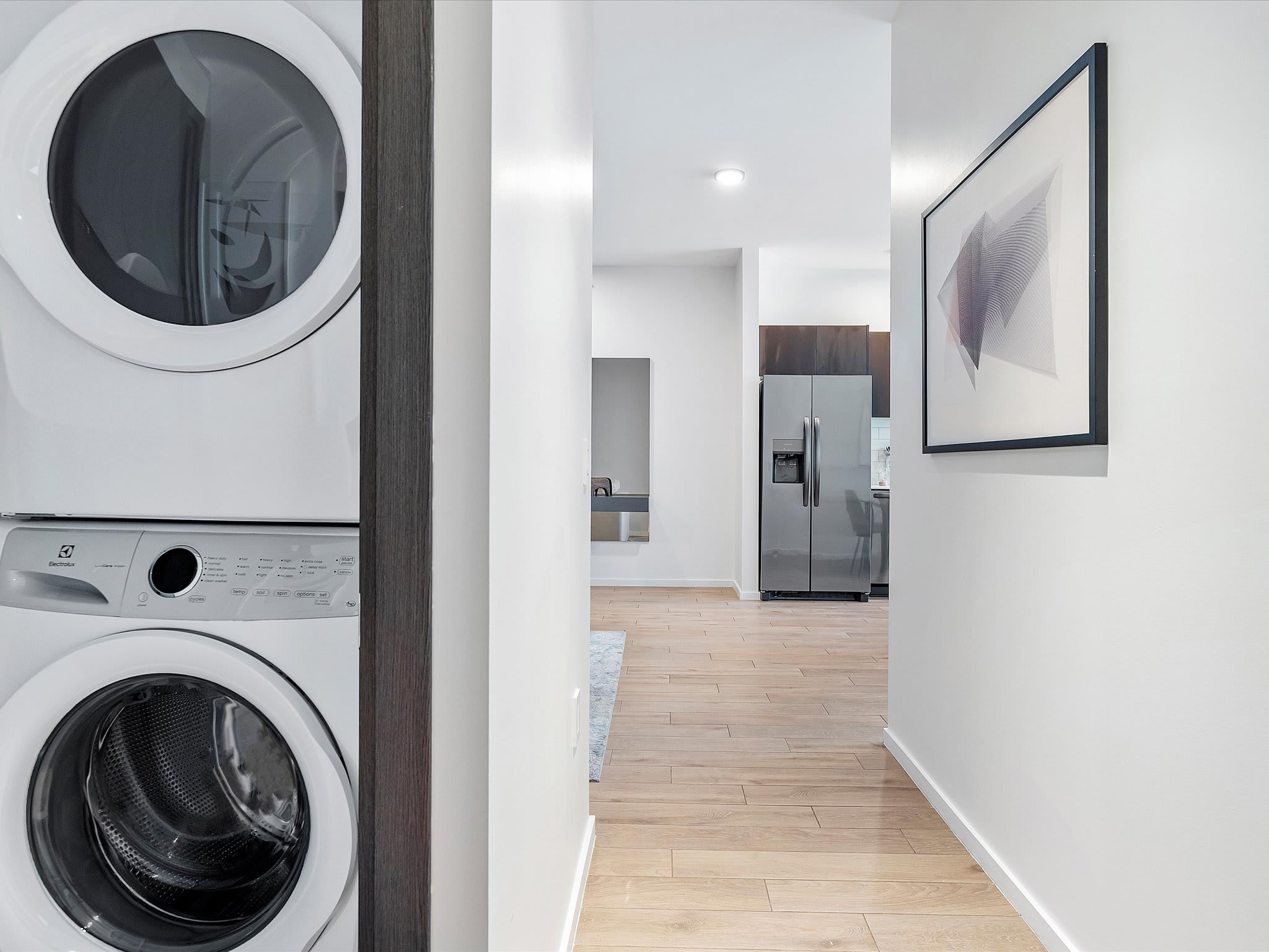 A modern hallway features light wood-look flooring and white walls, with a stacked white Electrolux washer and dryer unit visible on the left, and a view into a kitchen and living area further down the hall.