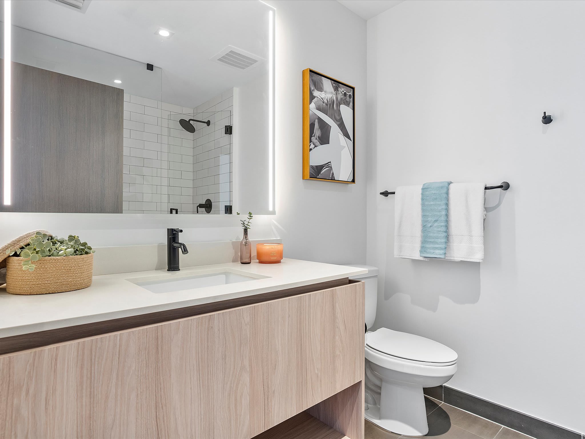 Modern bathroom with a light wood vanity, white countertop, black faucet, illuminated mirror, and a toilet, featuring white subway tile in the shower area and dark grey floor tiles.