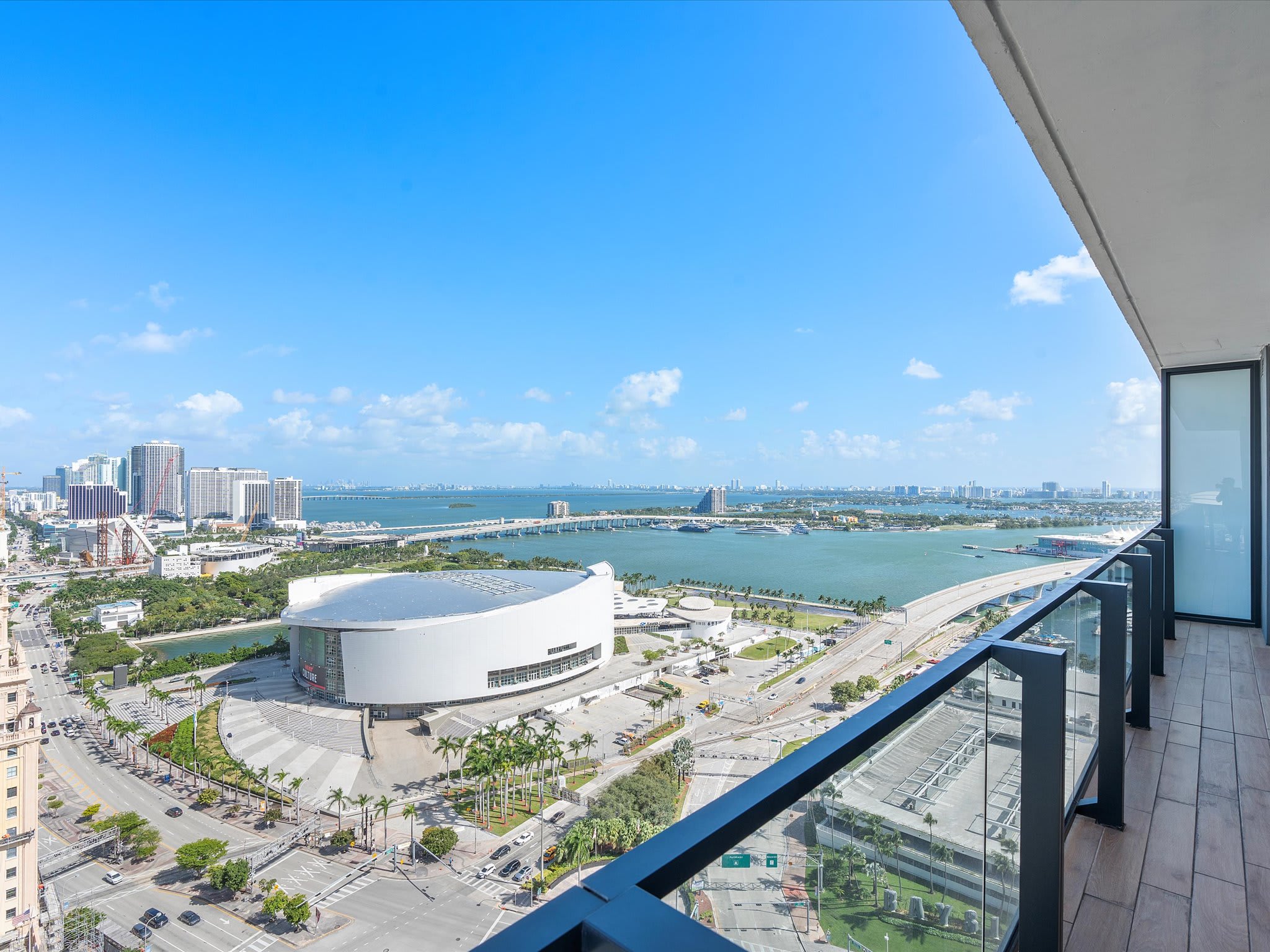 Expansive high-rise balcony with glass railings, offering breathtaking panoramic views of the city skyline, a large arena, and a bay with bridges under a clear blue sky.
