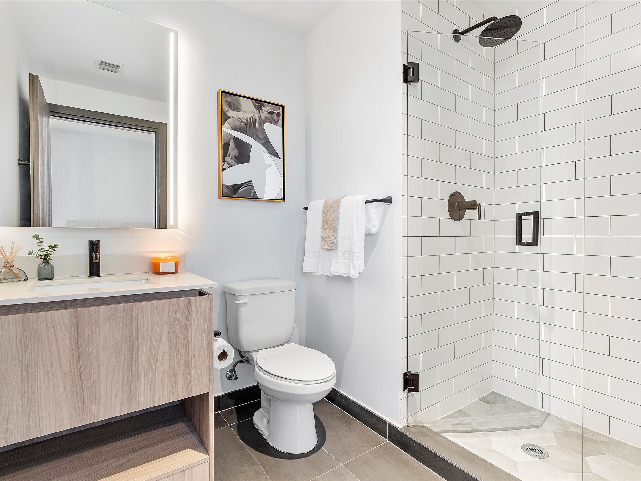 Modern bathroom with a light wood vanity, white countertop, black faucet, illuminated mirror, and a walk-in shower with white subway tiles and a glass enclosure, featuring dark grey floor tiles.