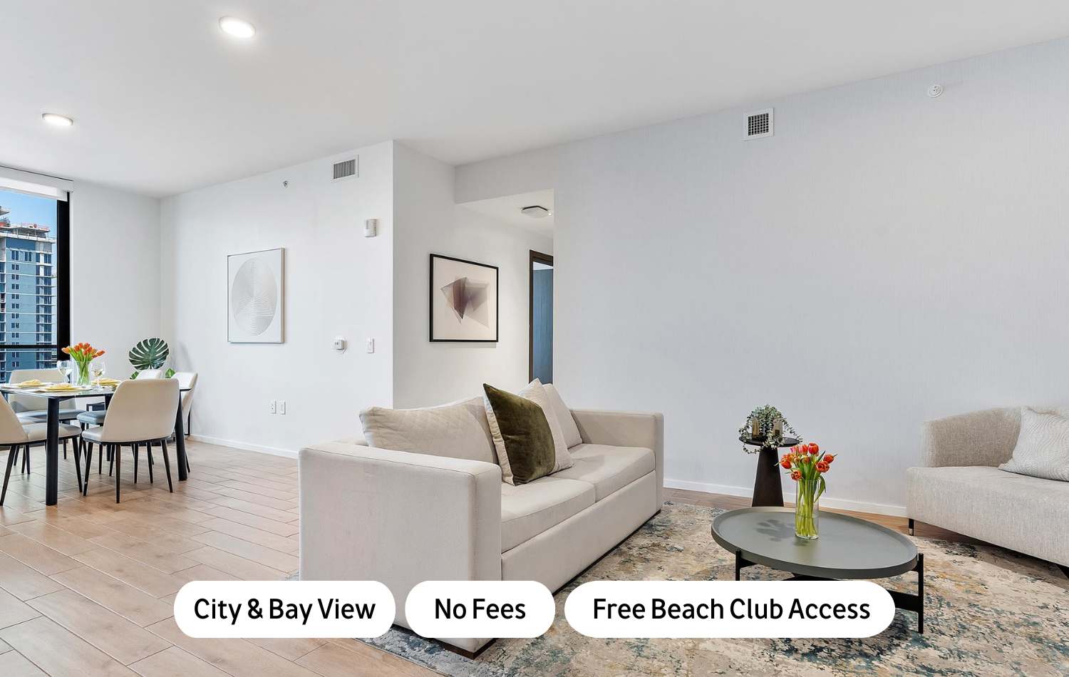 Bright open-concept living space with light wood flooring, a cream sofa with olive and dark green pillows, a round coffee table with flowers, and a dining area with a black marble table and light chairs, all under recessed lighting.