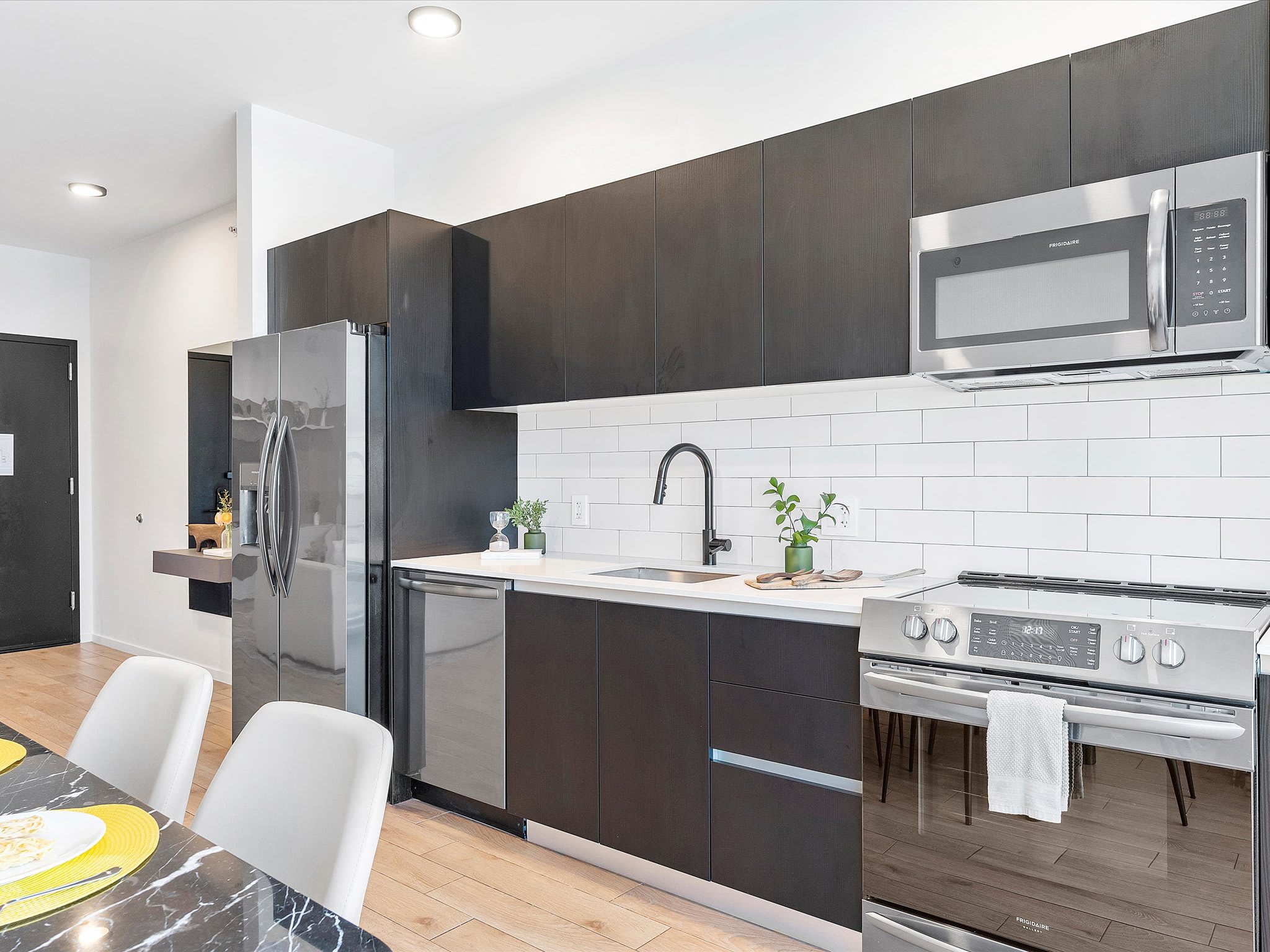 Contemporary kitchen featuring dark wood cabinetry, white subway tile backsplash, stainless steel refrigerator, dishwasher, microwave, and range, with a black gooseneck faucet over a sink, and light wood flooring.